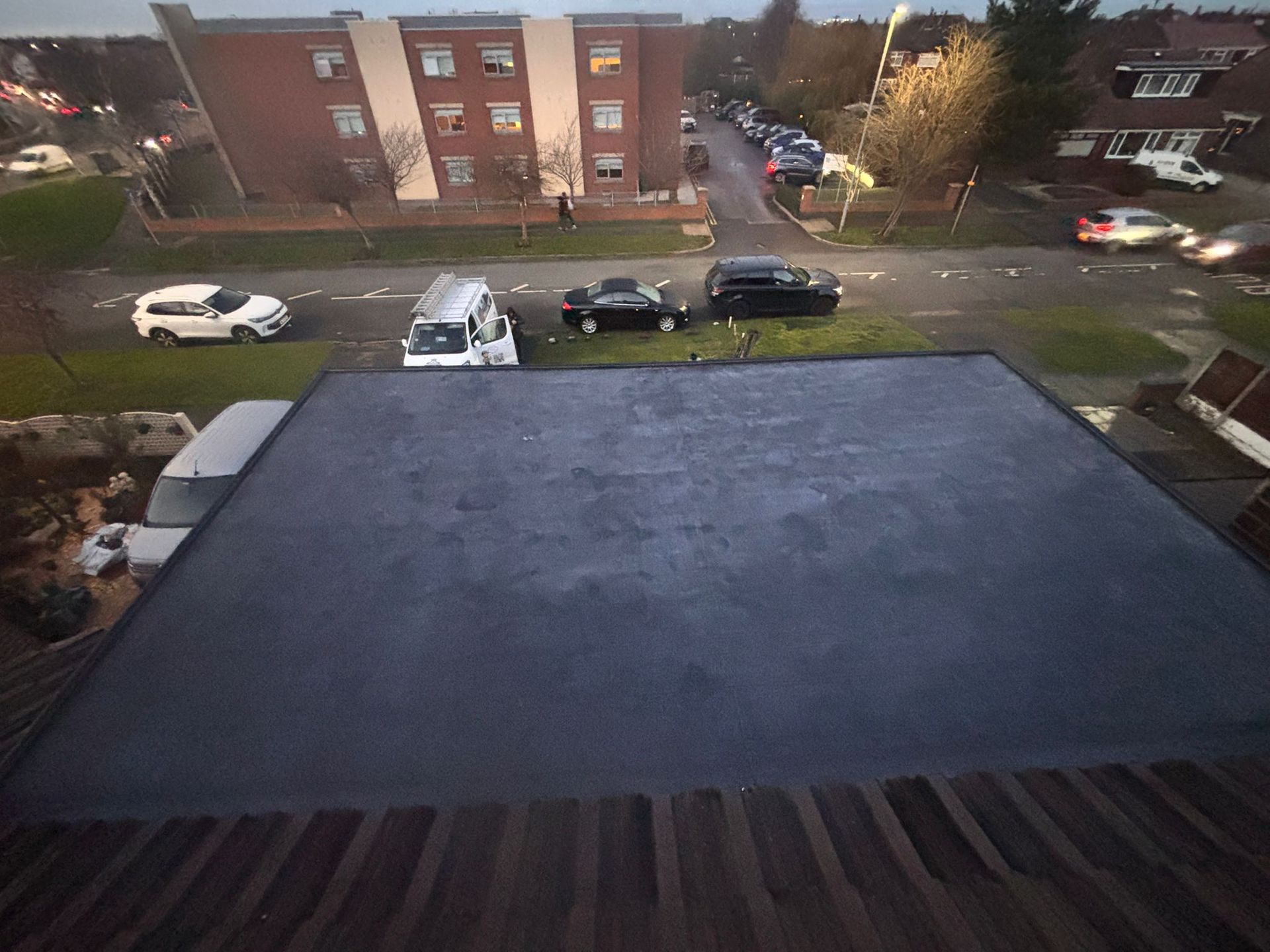 An elevated view of a flat, dark grey roof surface, with residential buildings and parked cars visible in the background.
