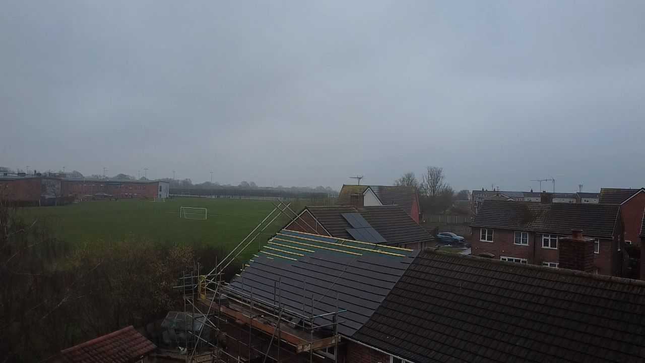 An elevated view of a house undergoing a roof replacement under a gray, overcast sky overlooking a green field.