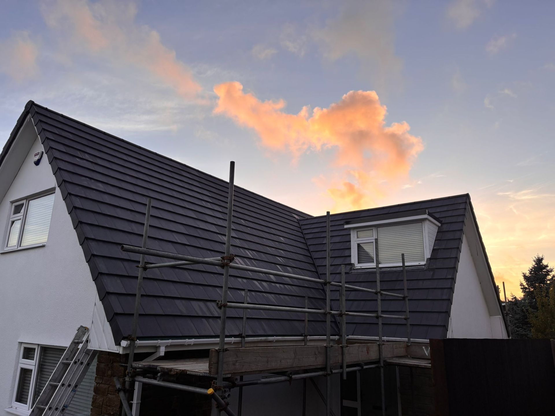 Scaffolding surrounds the roof and white exterior walls of a house at sunset, with dark grey roof tiles and two windows.
