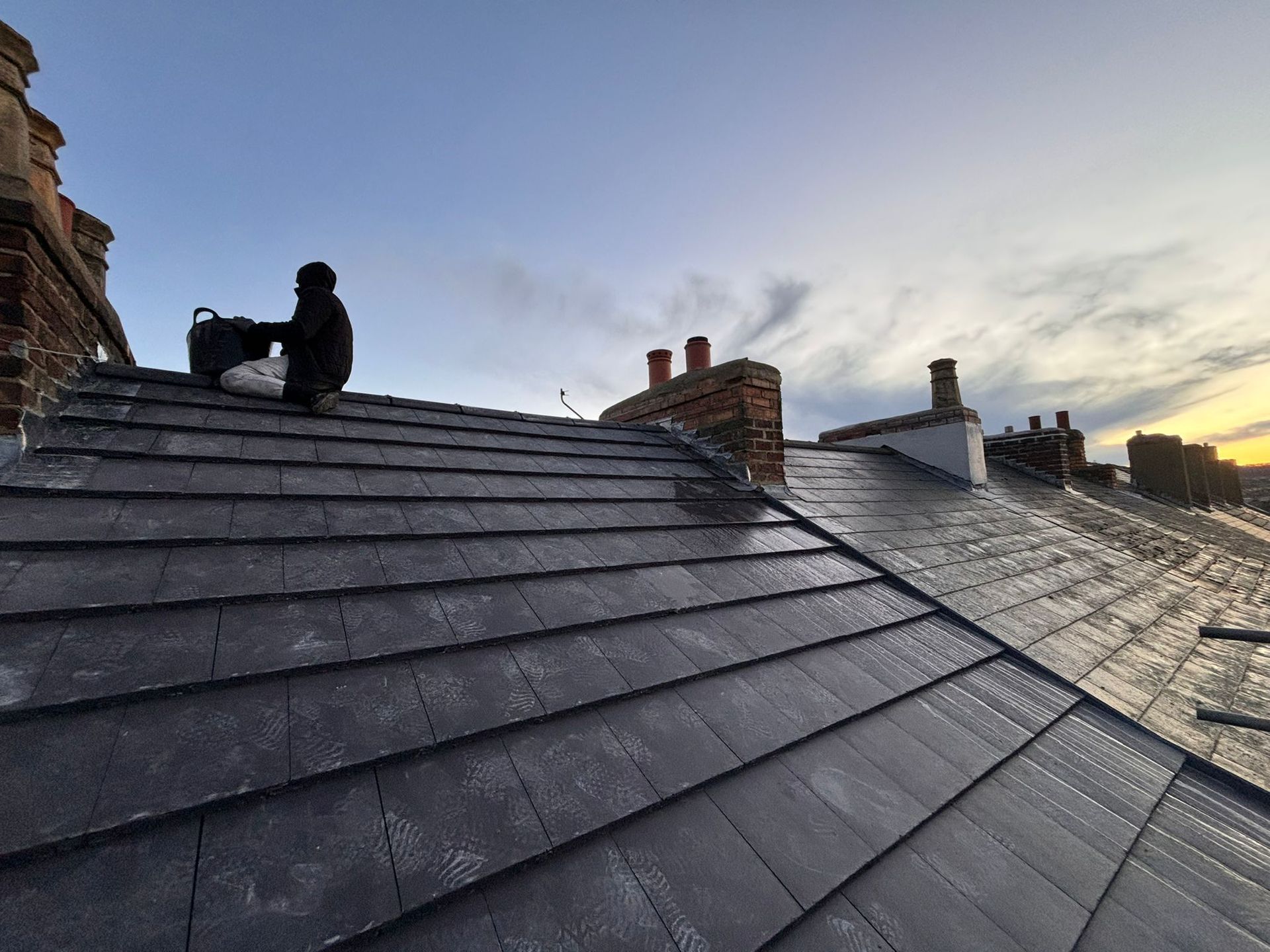 A person sits on a dark, slate-tiled pitched roof against a twilight sky with multiple brick chimneys visible.