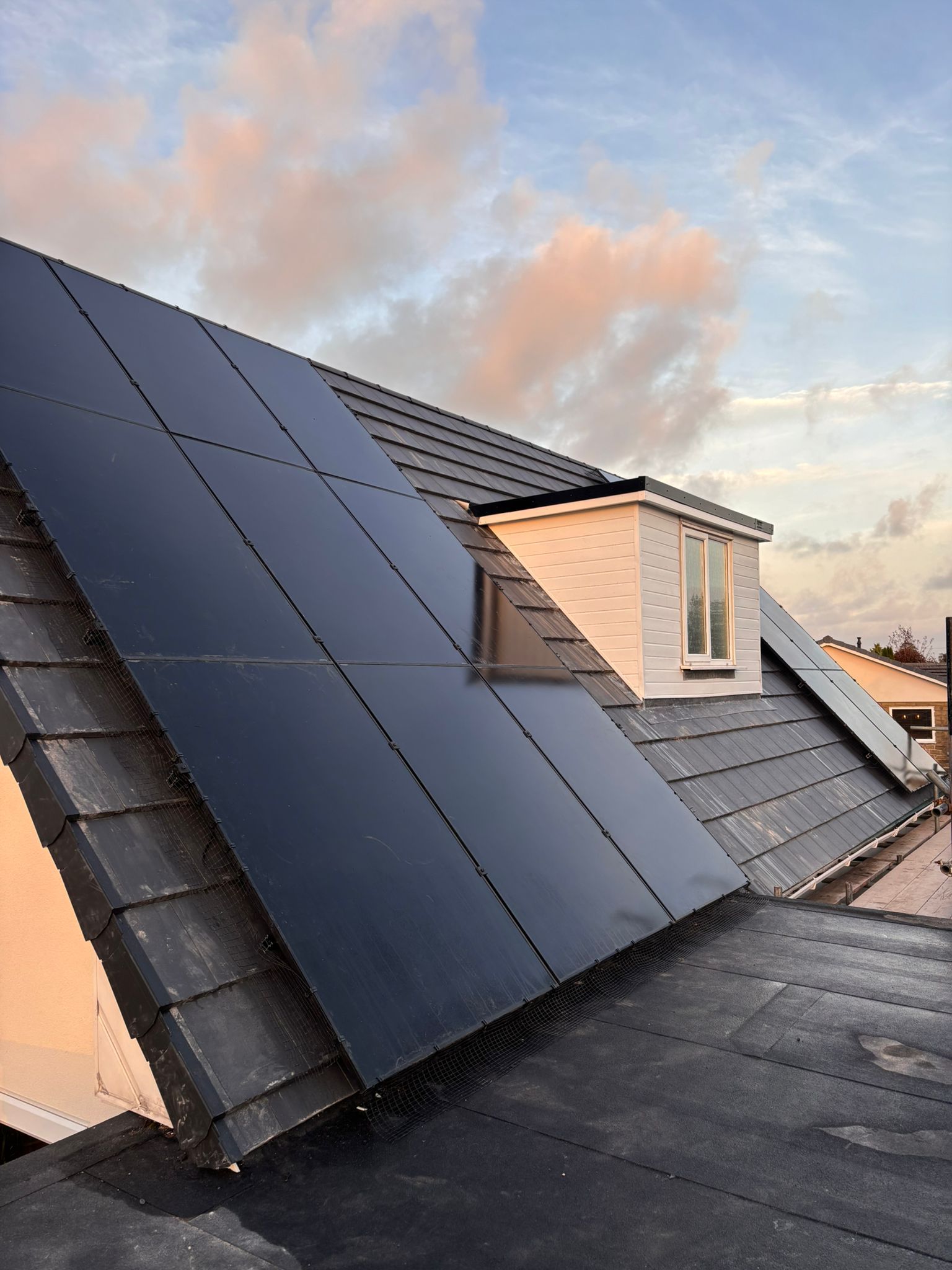 Sleek black solar panels installed on a residential dark-tiled roof next to a white dormer window at sunset.