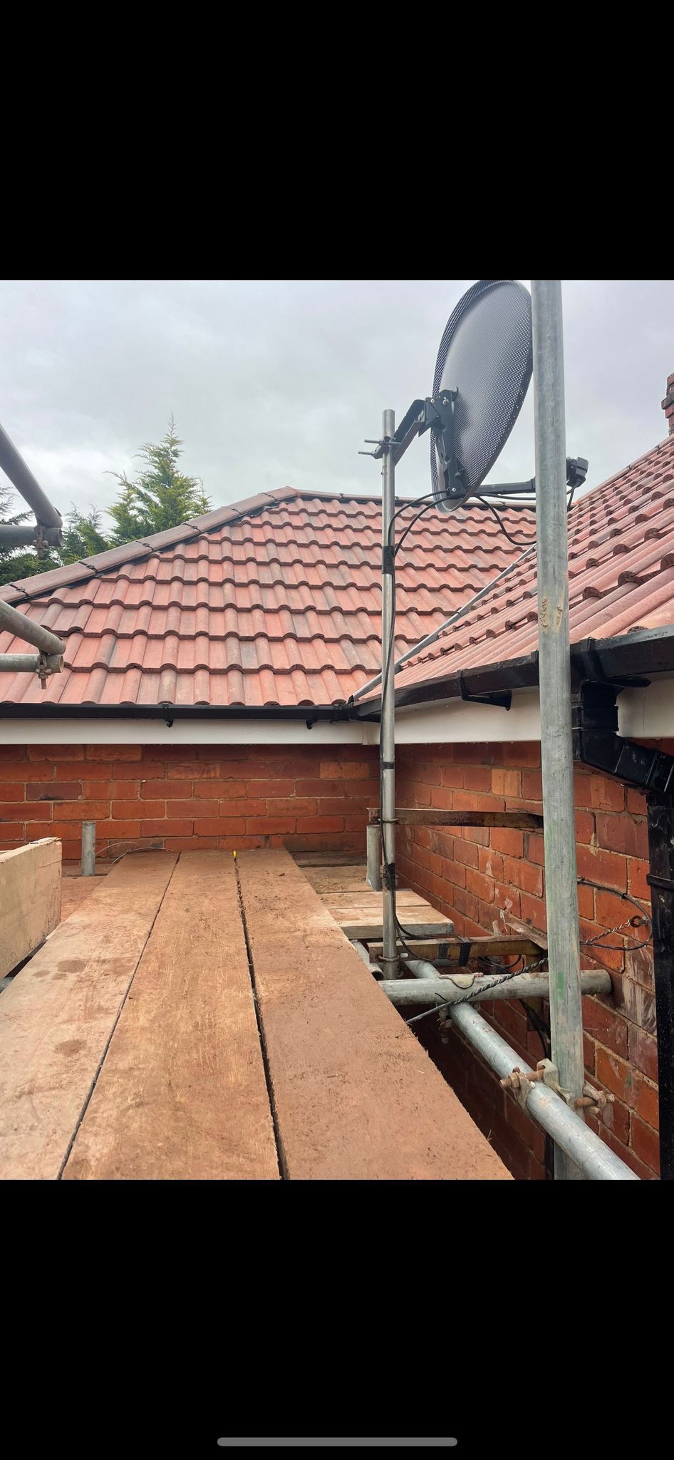 Scaffolding platform along a brick wall under a tiled roof, with a large circular saw blade attached to an upright pole.