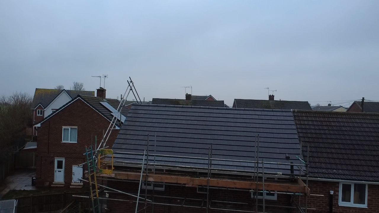A house roof under construction, featuring new dark tiles, partially finished scaffolding, and surrounding homes.