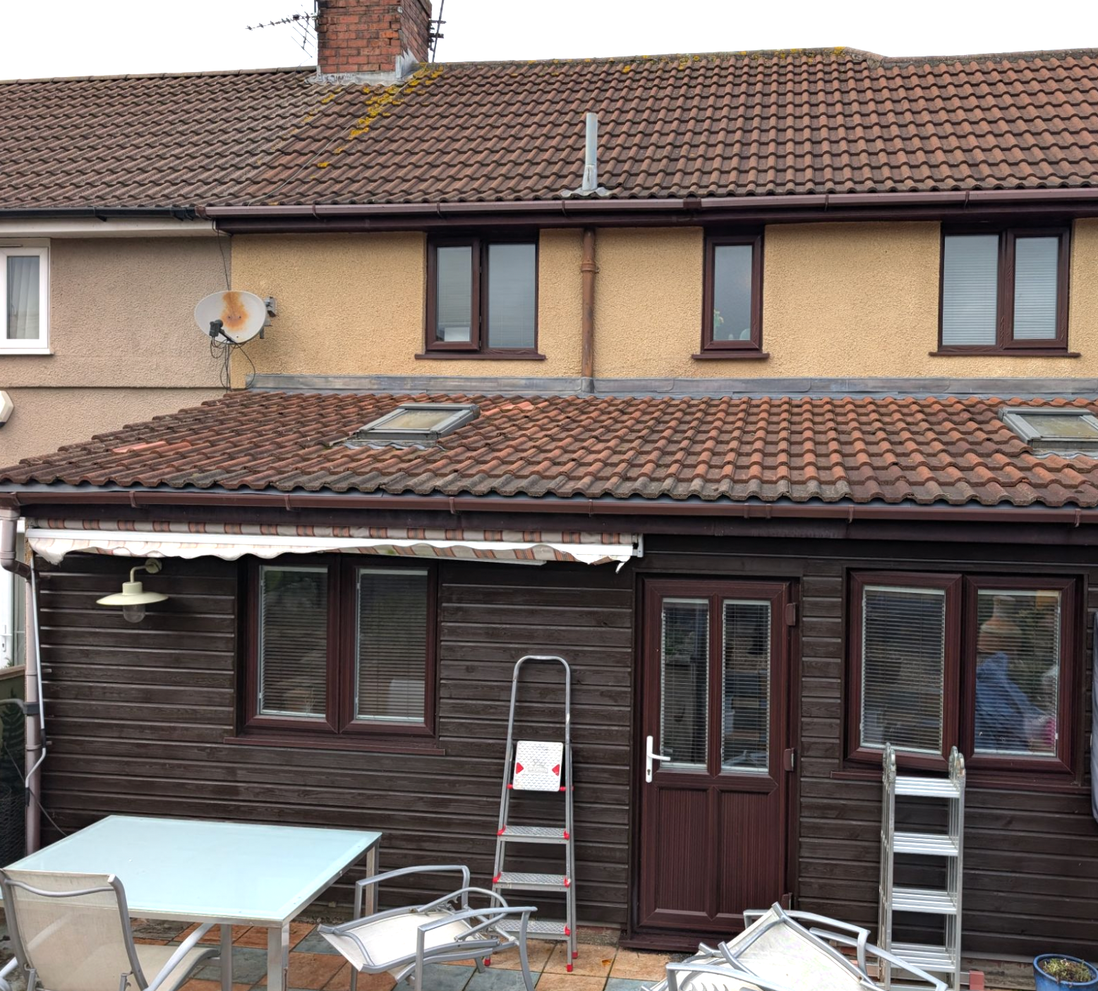 Backyard patio with a dark wood conservatory, ladders, and scattered renovation materials beneath a tan house.