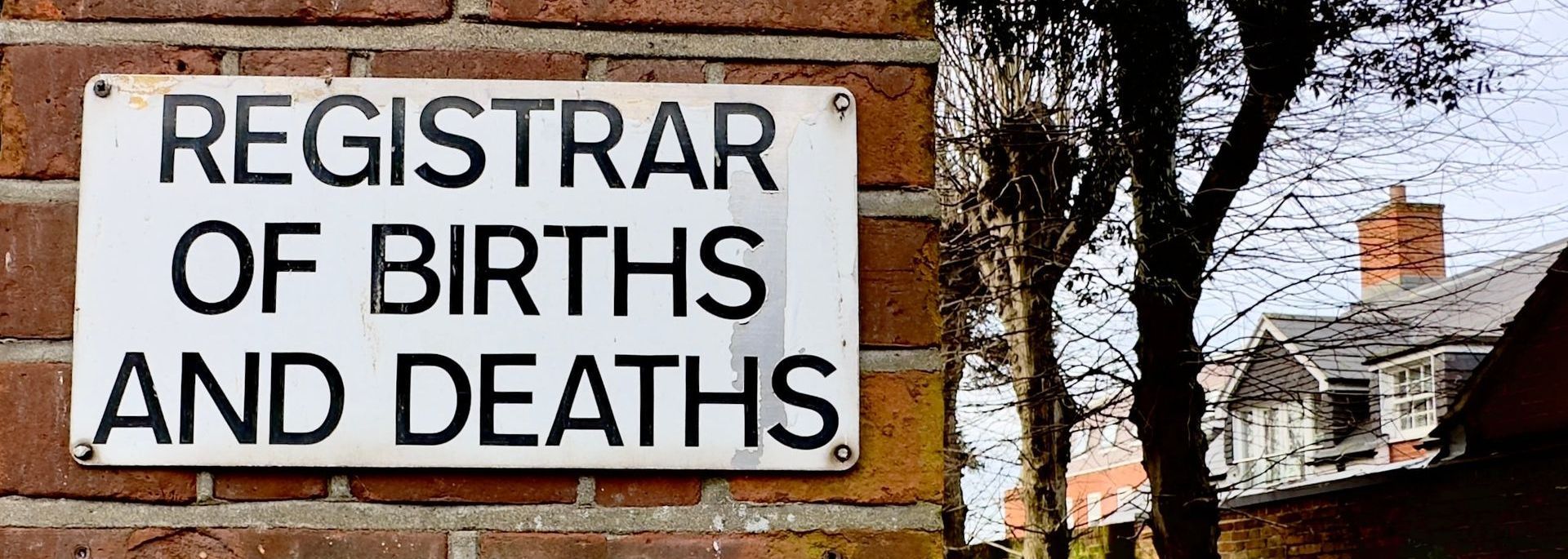 A closeup of a sign for the Registrar of Births and Deaths on a brick wall in the UK.