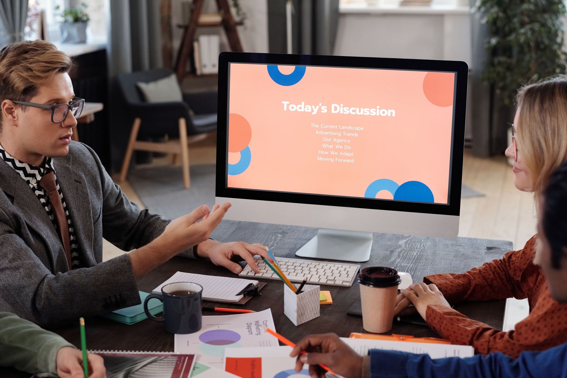 Two coworkers in a meeting room discuss a presentation on a laptop screen.