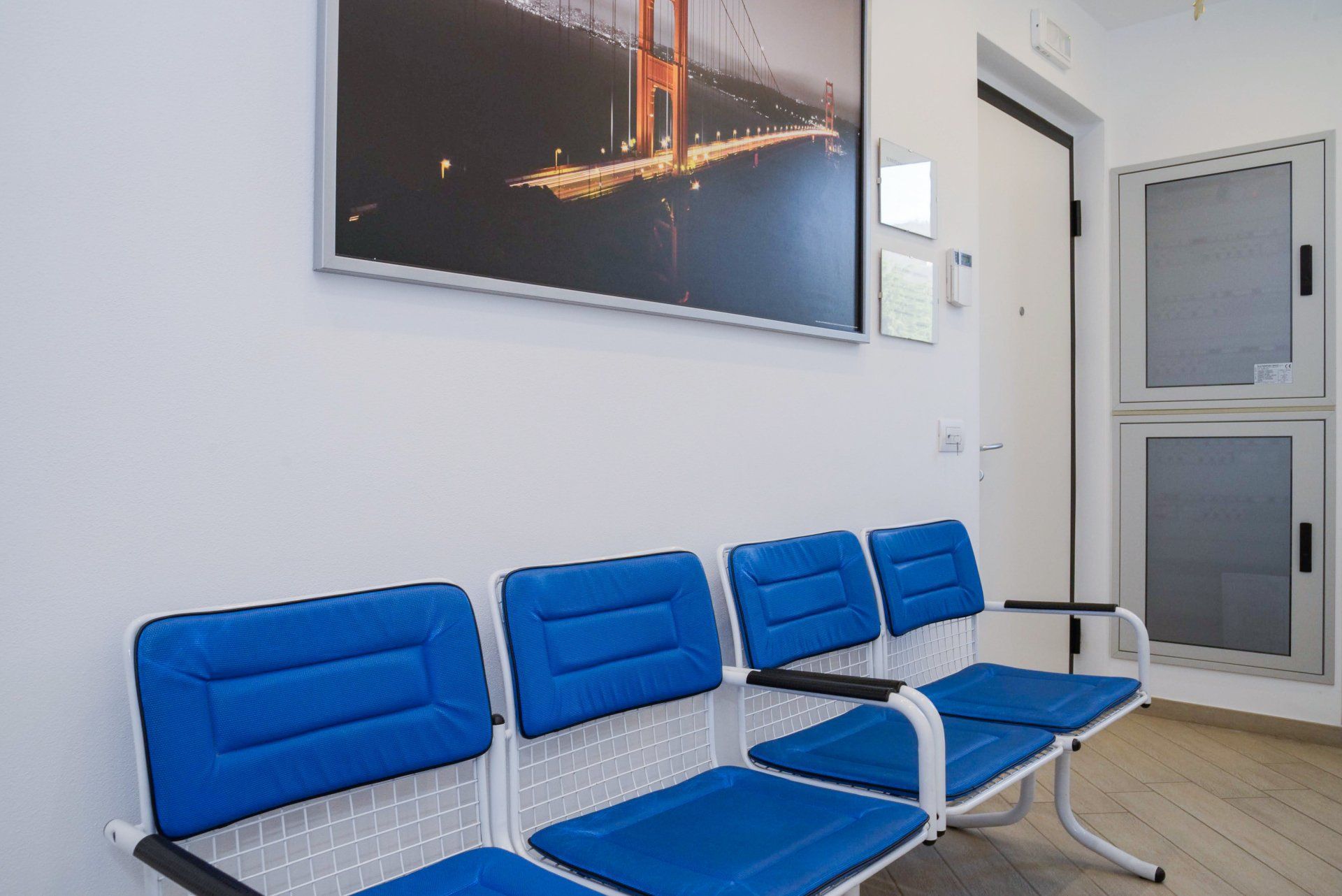 Sedie d'attesa blu in una hall luminosa con opere d'arte alle pareti e una porta sullo sfondo.
