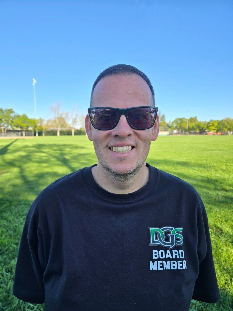 Football coach with arms crossed, looking stern on a sunny field, players in the background.