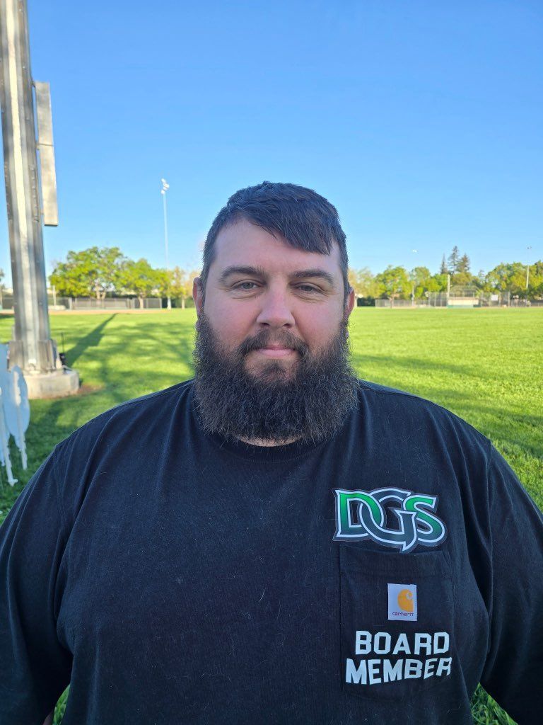 Man in green polo stands on a football field, arms crossed, stadium in background.