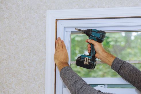 Hands installing a white window frame with a cordless drill.