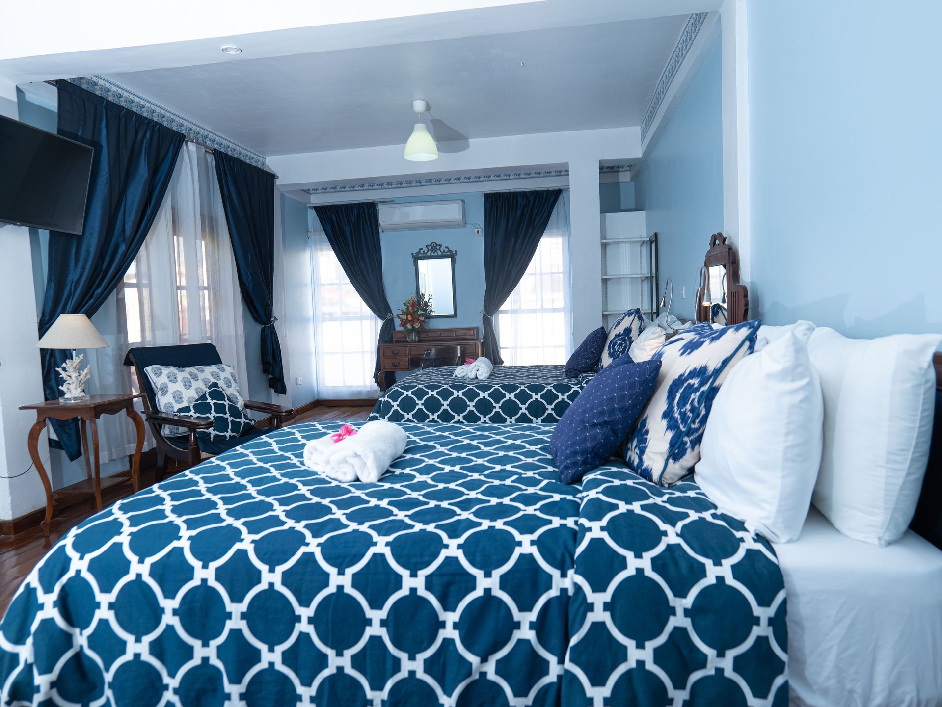 A bedroom with two beds featuring blue and white geometric bedding, dark curtains, and light blue walls.