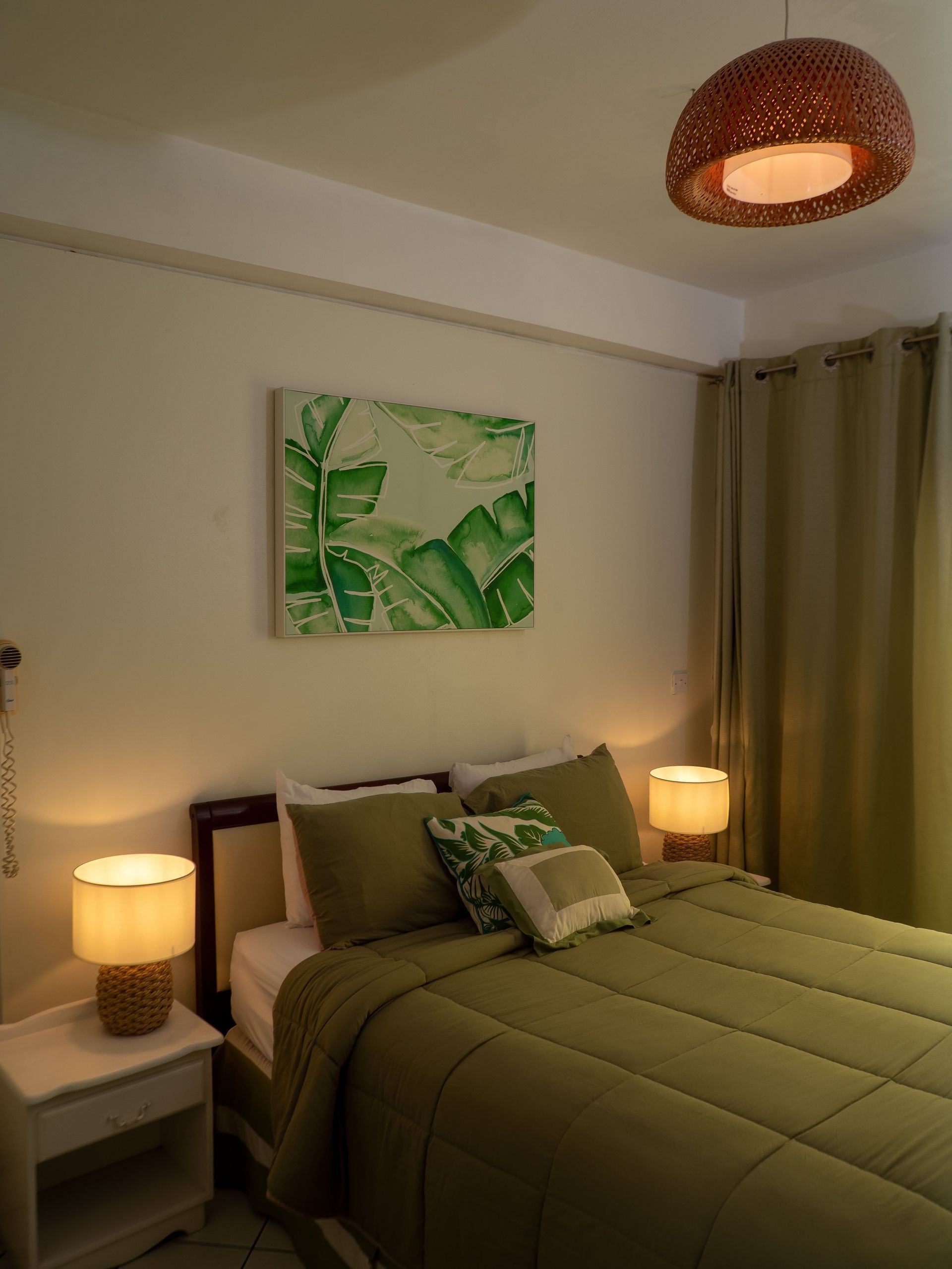 A cozy bedroom with olive green bedding, a botanical wall print, warm bedside lamps, and a woven overhead pendant light.