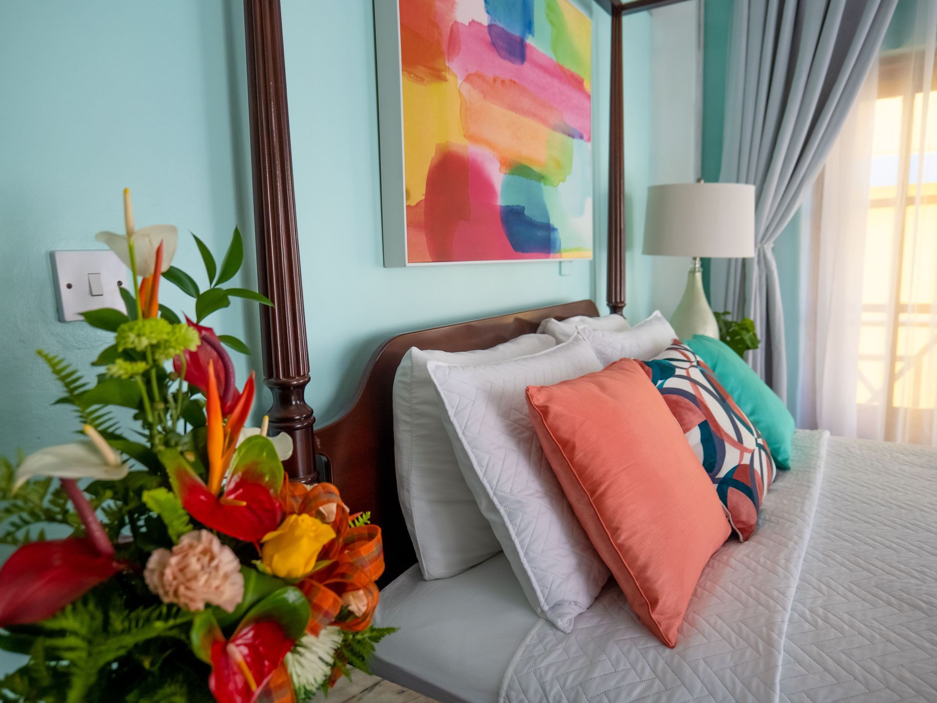 A tropical flower bouquet in the foreground of a bedroom with light blue walls, a four-poster bed, and abstract art.