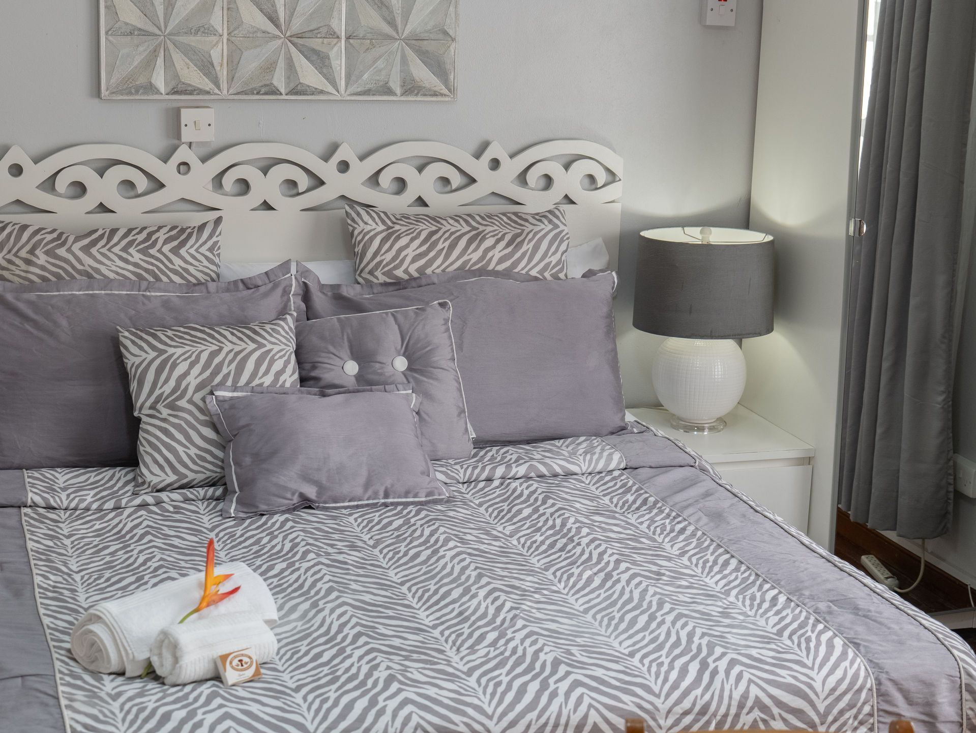 A bedroom with a white ornate headboard, grey bedding with zebra print accents, and a lamp on a bedside table.