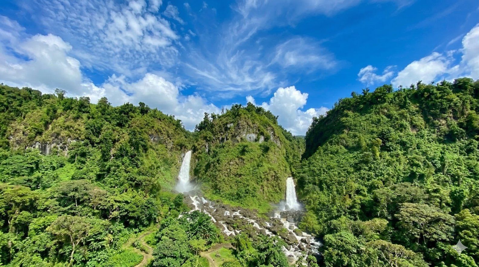 A panoramic view of two cascading waterfalls nestled between lush, green, forested mountains under a bright blue sky.