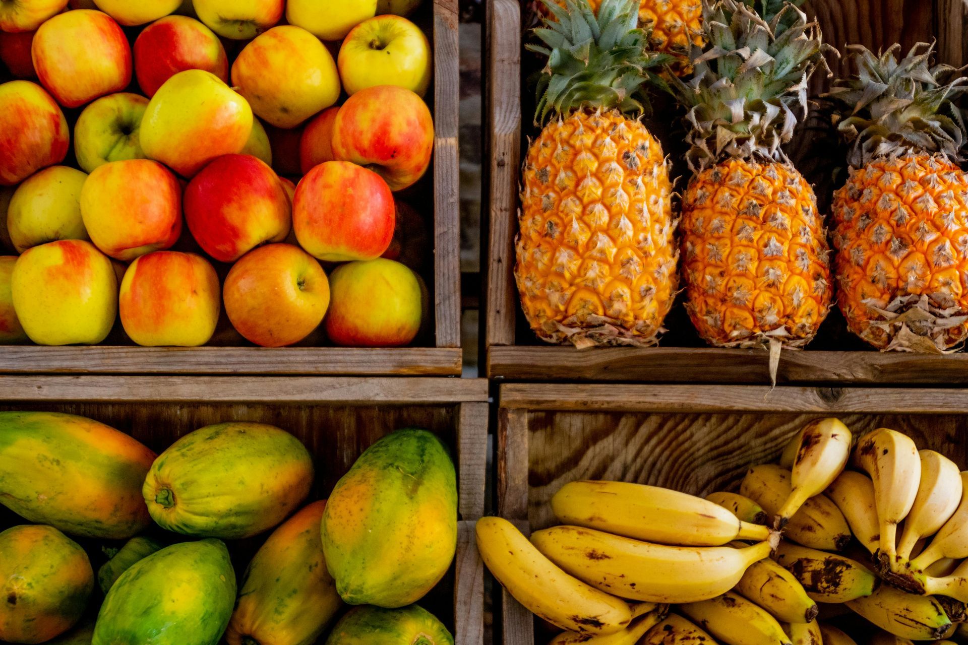 Four wooden crates filled with apples, pineapples, papayas, and bananas displayed at a market.
