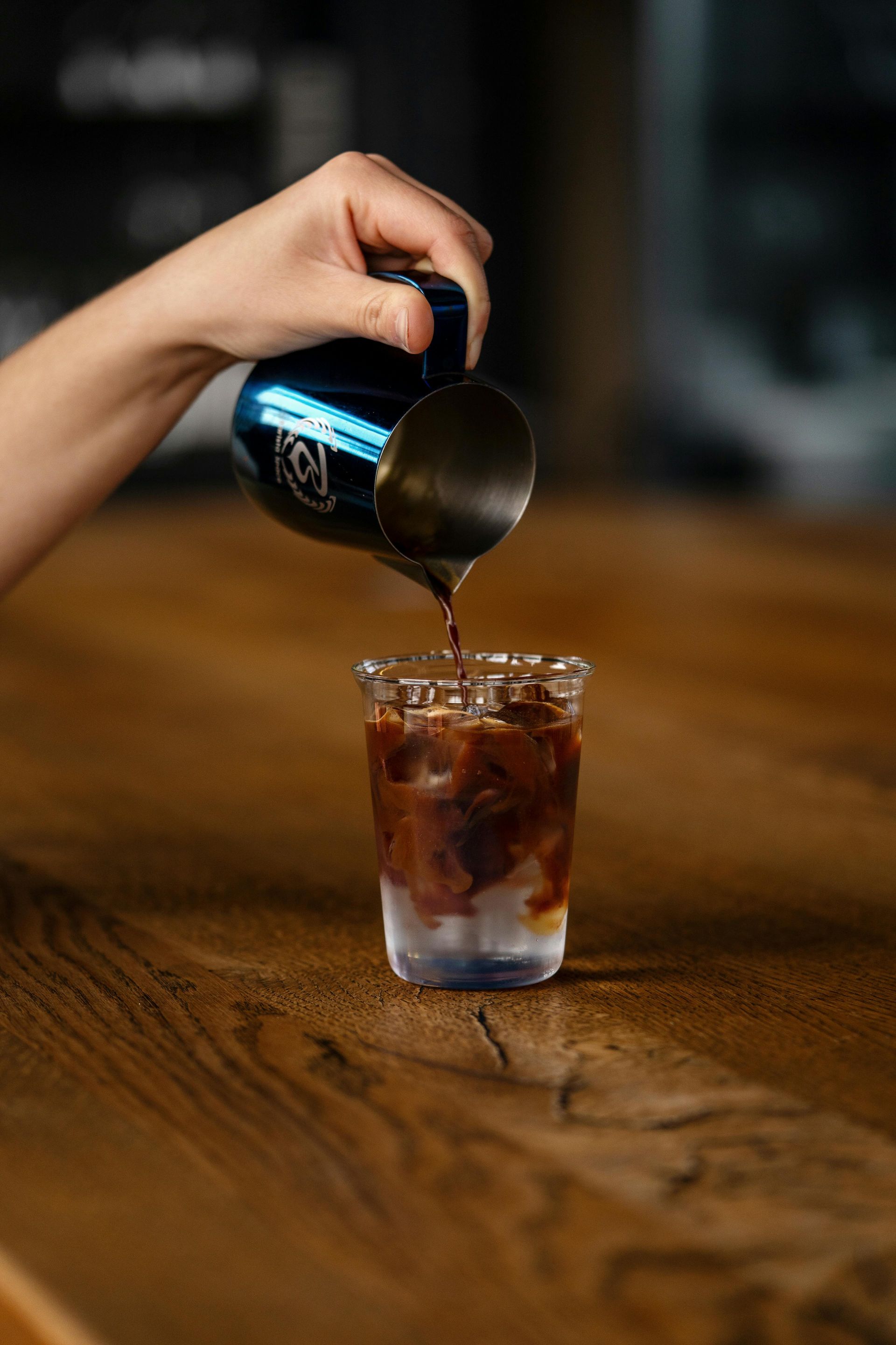 A hand pours espresso from a blue metal pitcher into a glass filled with ice and milk on a wooden counter.