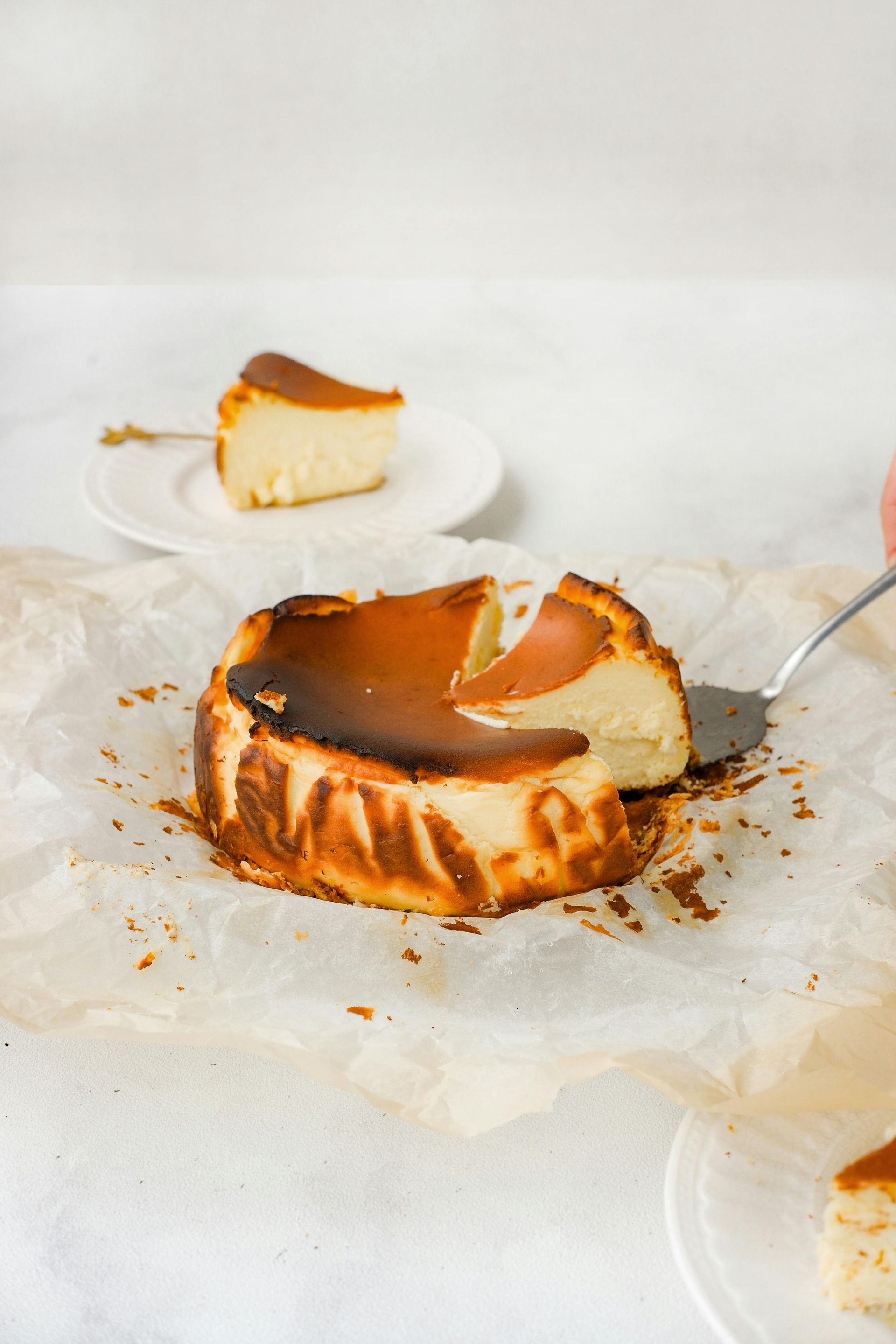 A sliced Basque burnt cheesecake on parchment paper, with a serving being lifted next to a small plate with another slice.