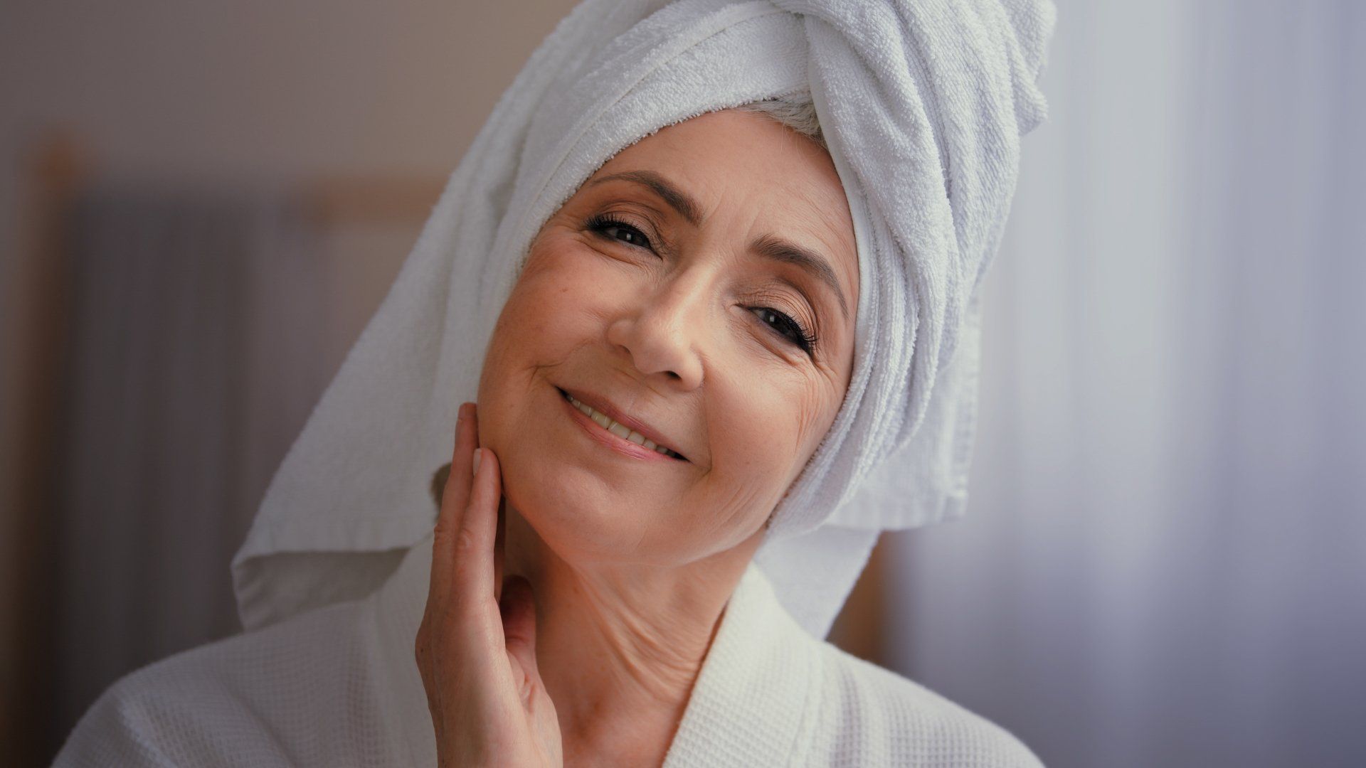 A woman with a towel wrapped around her head is smiling and touching her face.