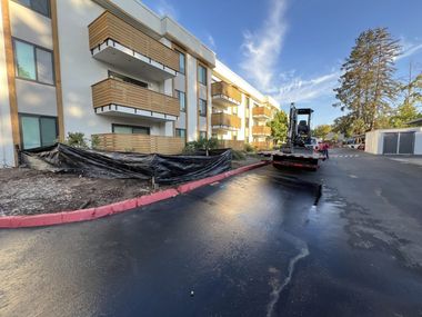 Damaged curb and burned landscaping beside an apartment building, with a skid steer on wet pavement