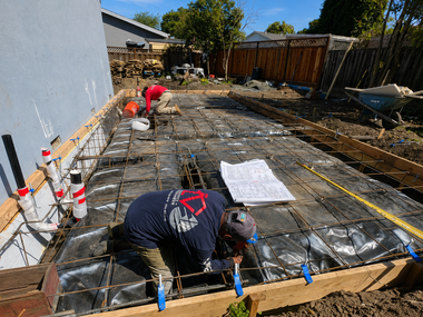 Workers laying rebar and concrete forms for a patio slab in a backyard under construction