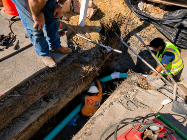 Utility workers repairing a broken pipe in an excavation trench with exposed dirt and tools.