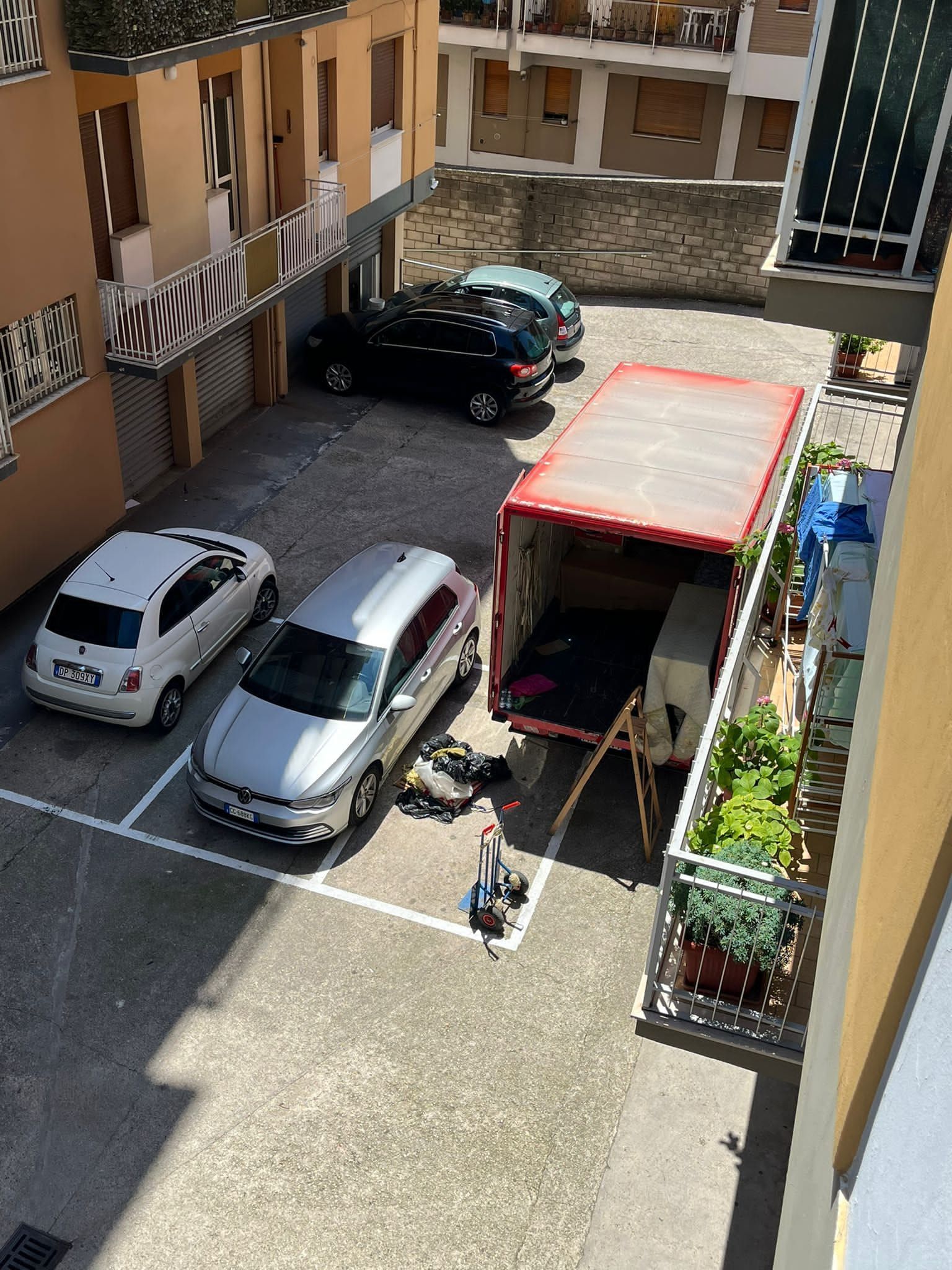 Auto parcheggiate in un cortile; un camion in movimento viene caricato, si vedono piante ed edifici.