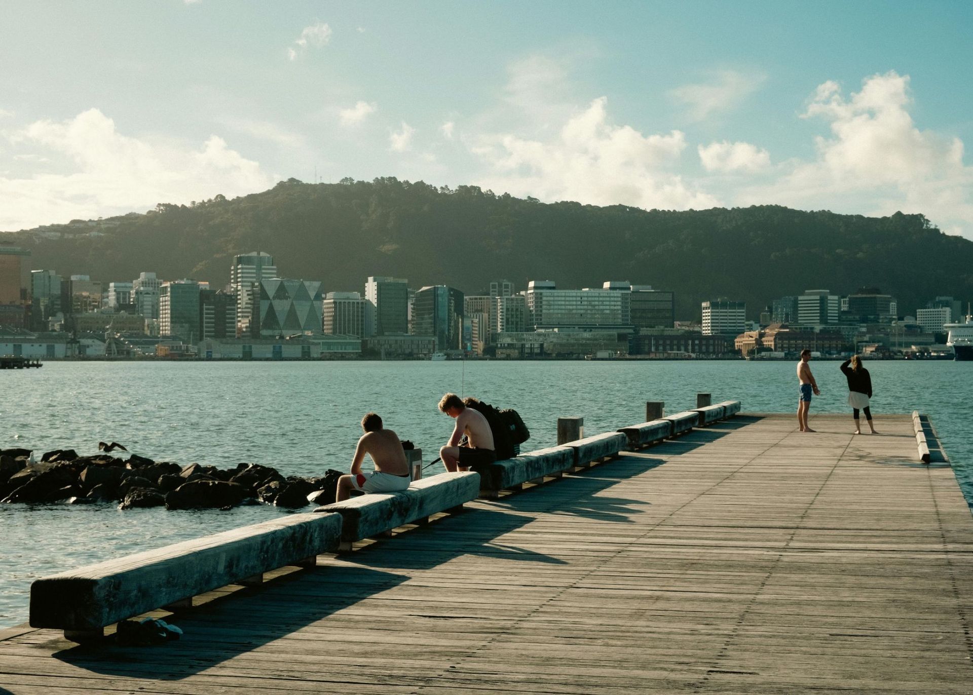 People sitting on a pier by the water with a city skyline and hill in the background.