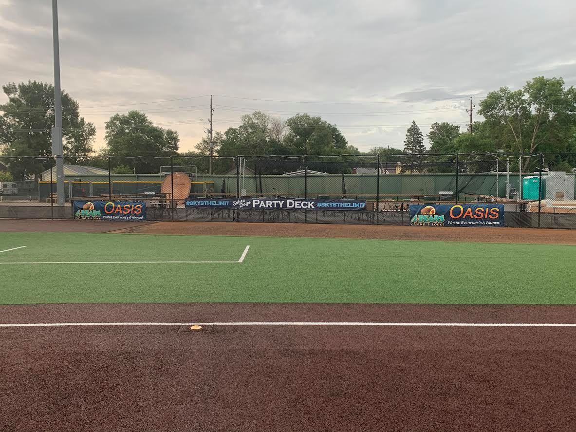 Baseball field with green turf, brown infield, and players near the dugout under a cloudy sky