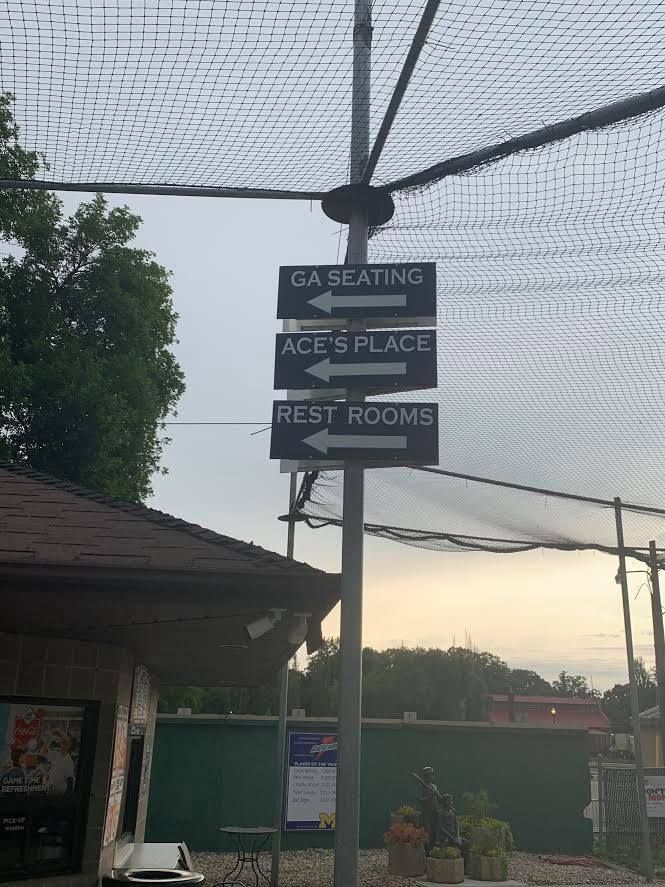 Outdoor sign for a driving range and golf shop, with arrows for ace's place and rest rooms under a netted canopy