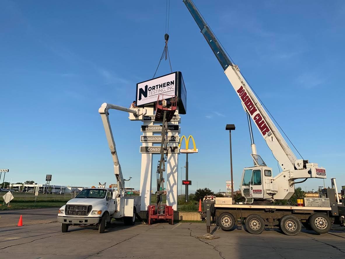 Crane lifting a roadside sign beside a truck and van on a sunny roadway.