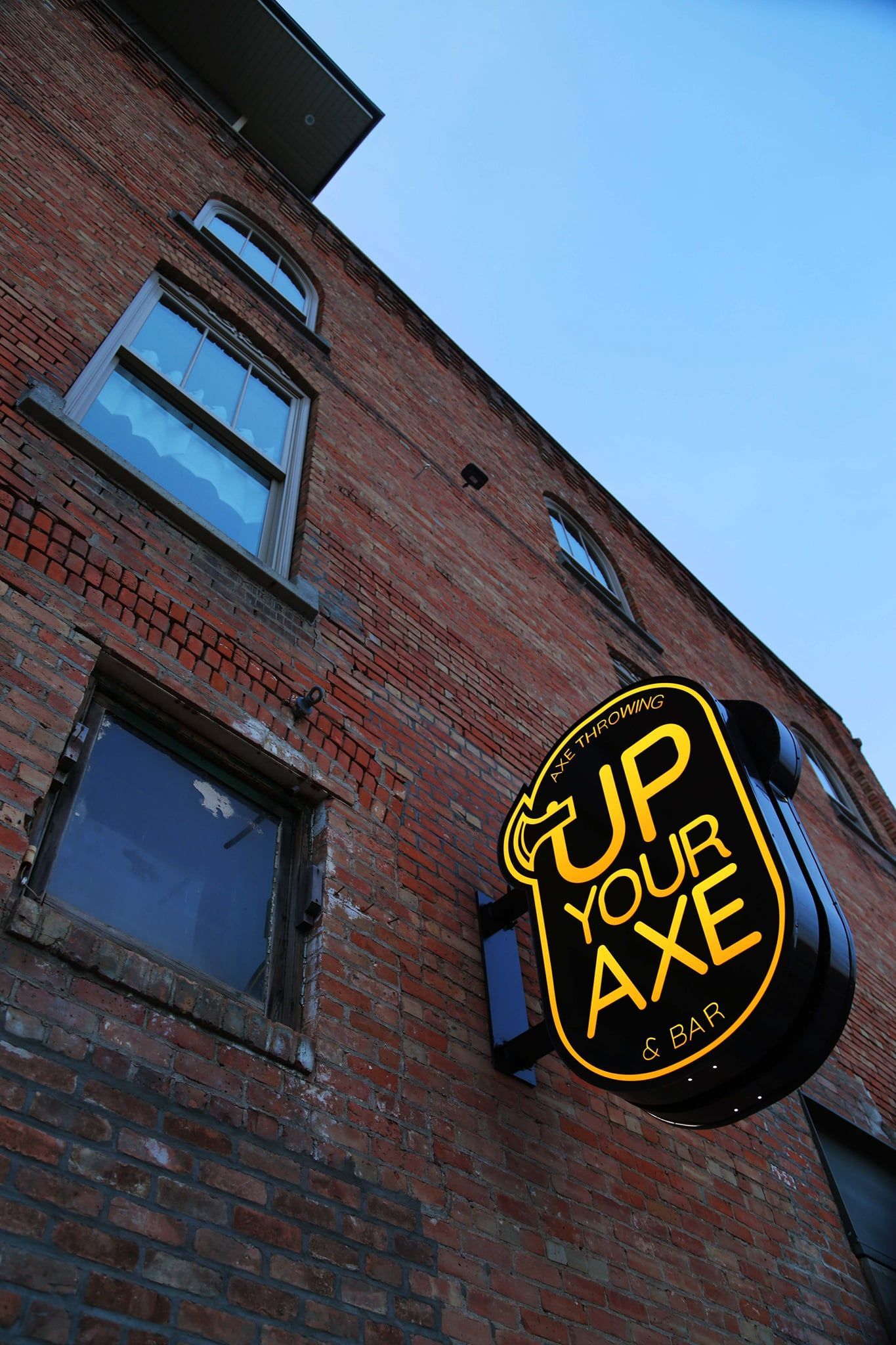 Brick building with a round black-and-yellow neon sign reading “UP YOUR AXE” under a blue sky