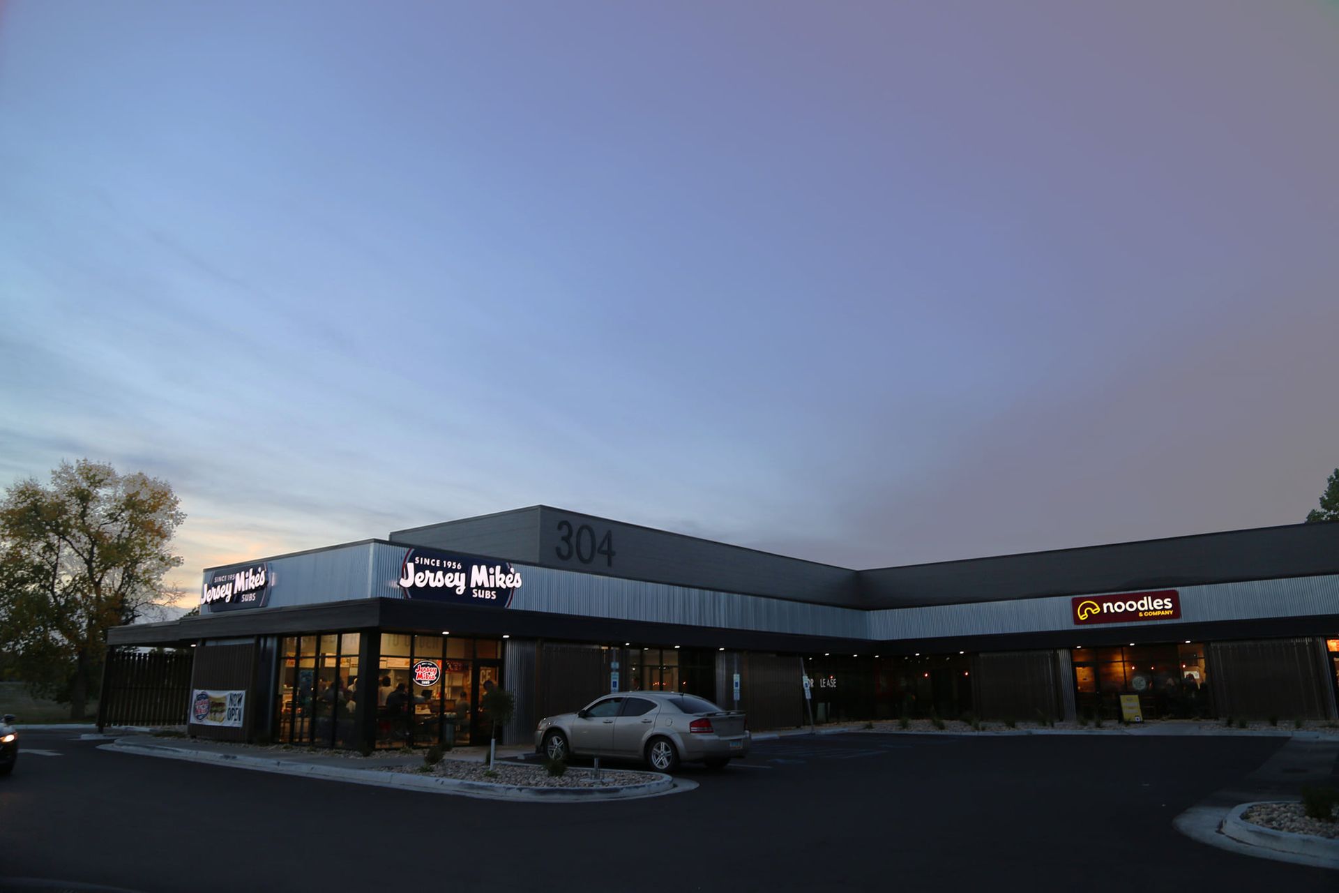 Shopping center at dusk with storefronts lit and a car parked in front.