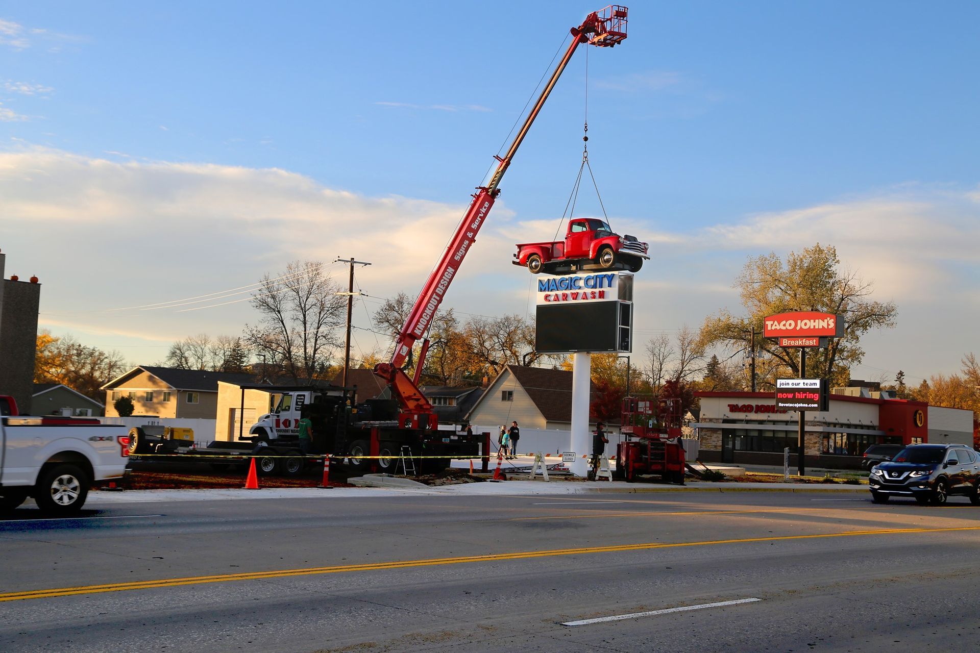 Crane lifting a large digital sign beside a road at dusk, with traffic and parked cars nearby