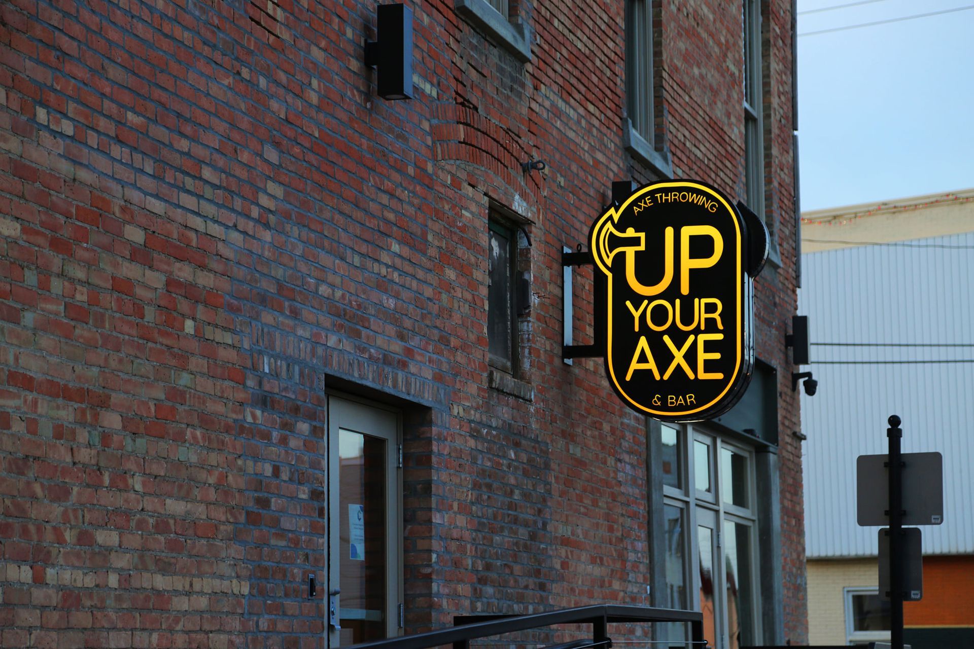Round neon sign on brick building reads “UP YOUR AXE” in yellow and black.