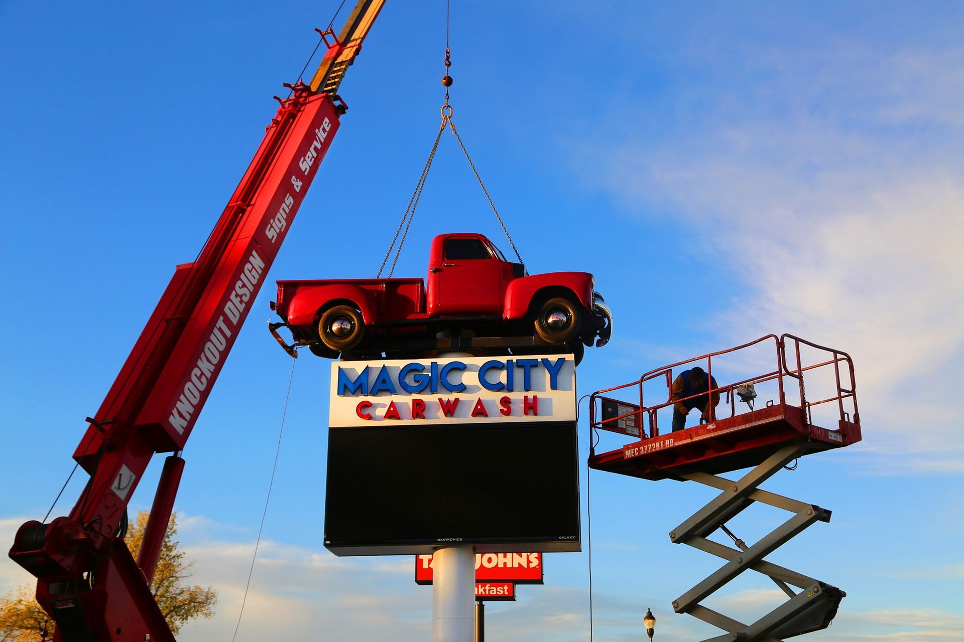 Red tow truck lifted by crane above a Magic City sign with a scissor lift beside it