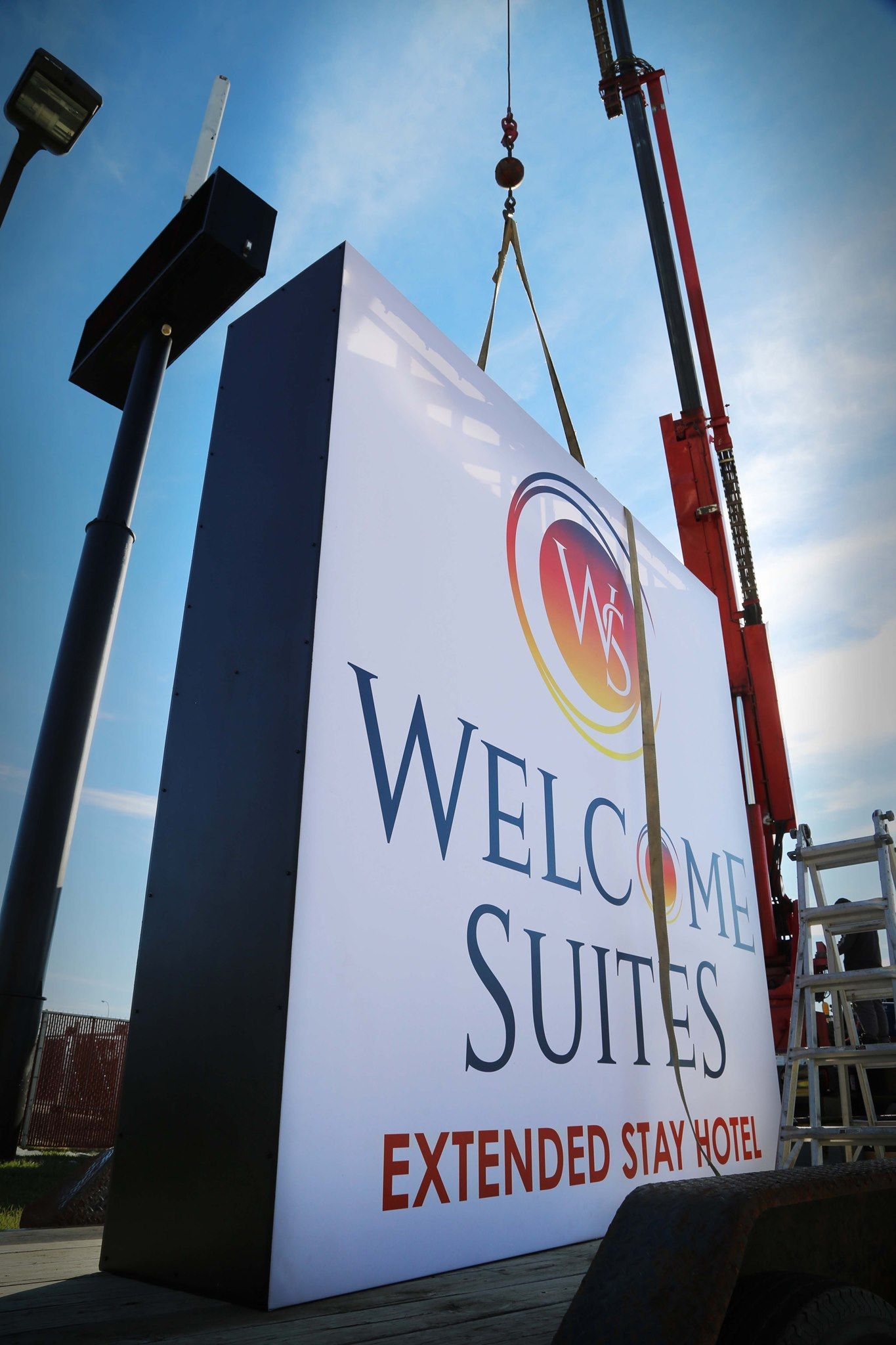 Tall hotel sign reading “Welcome Suites Extended Stay” against a blue sky, being hoisted by cranes