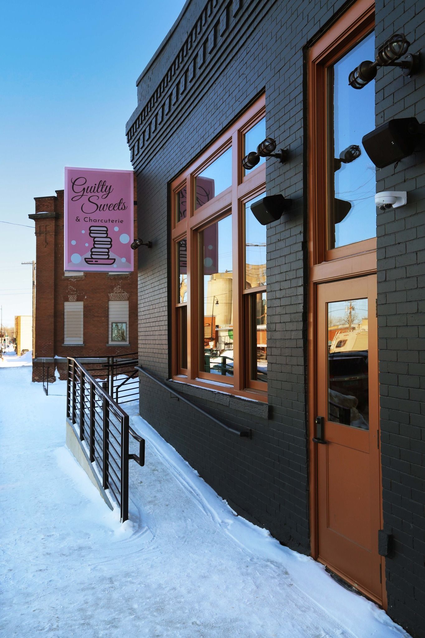 Snowy sidewalk beside a dark brick building with orange-framed windows and a pink hanging sign