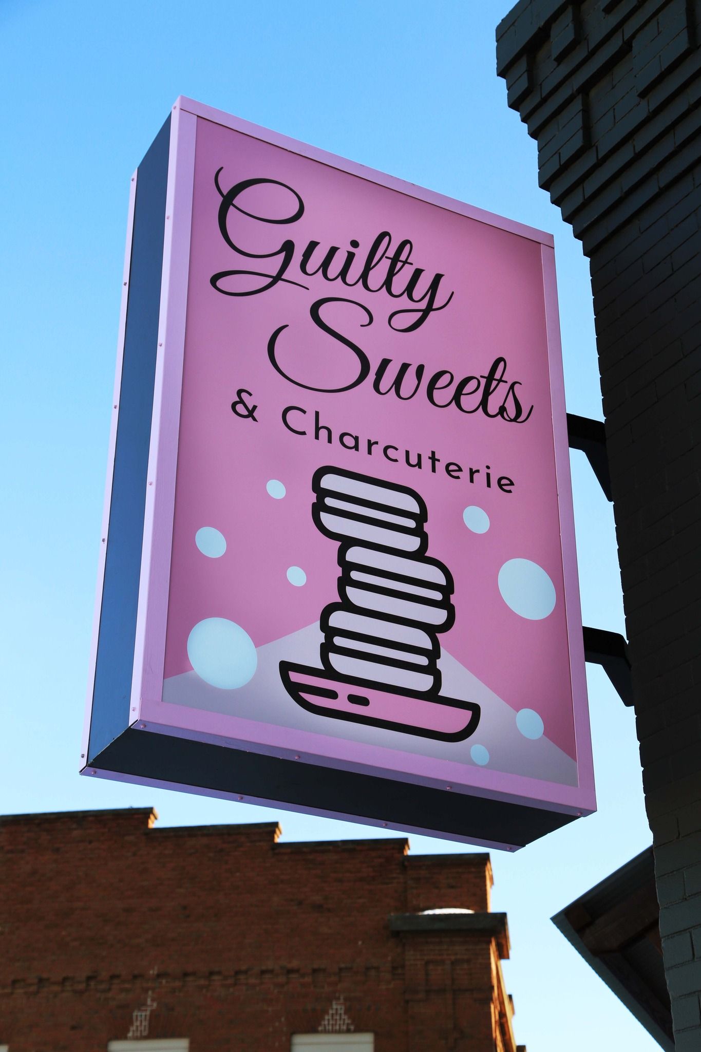 Pink storefront sign for “Guilty Sweets & Charcuterie” with stacked dessert graphic on a building exterior