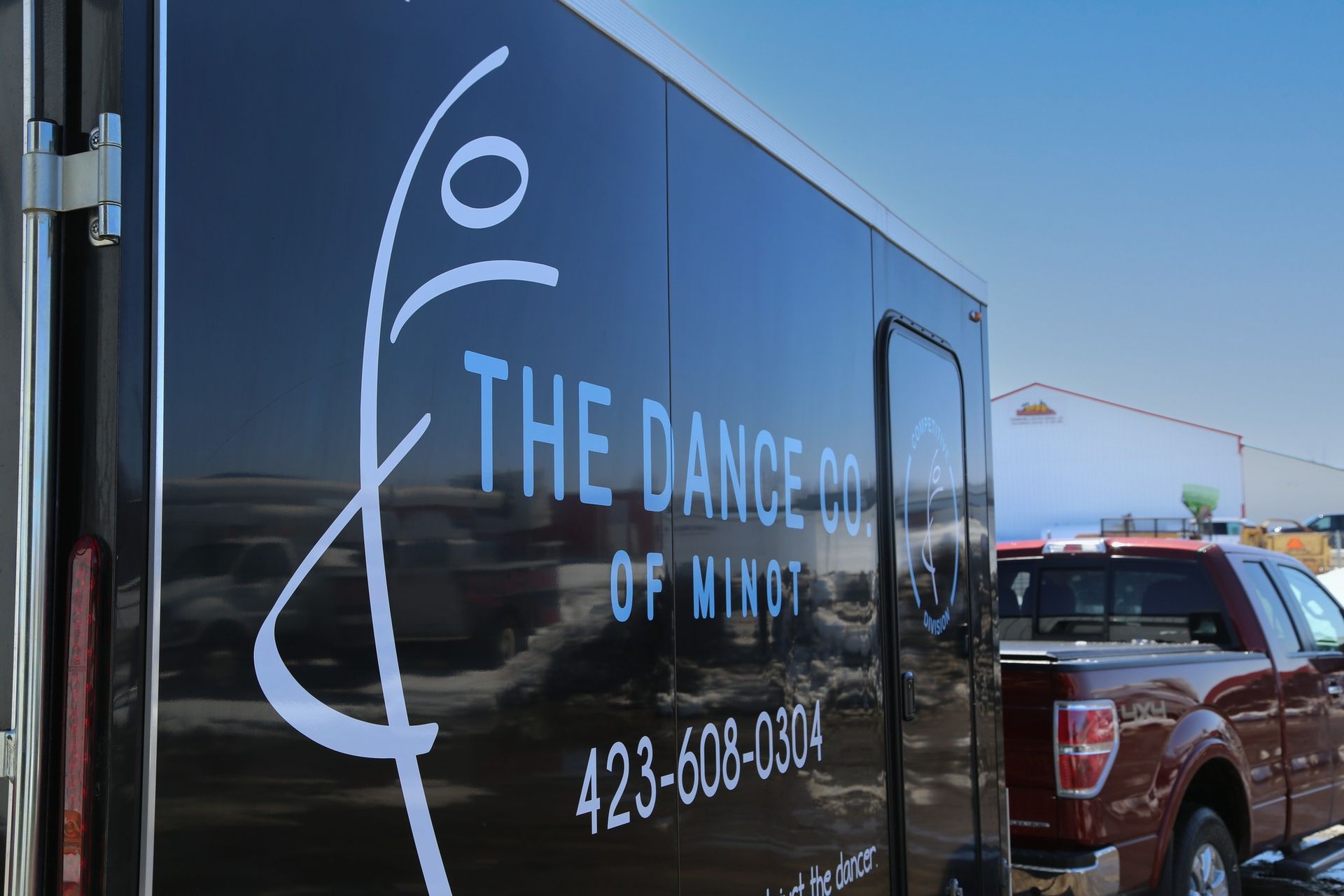 Black trailer with “The Dance of Motion” logo and phone number parked outdoors near a red truck.
