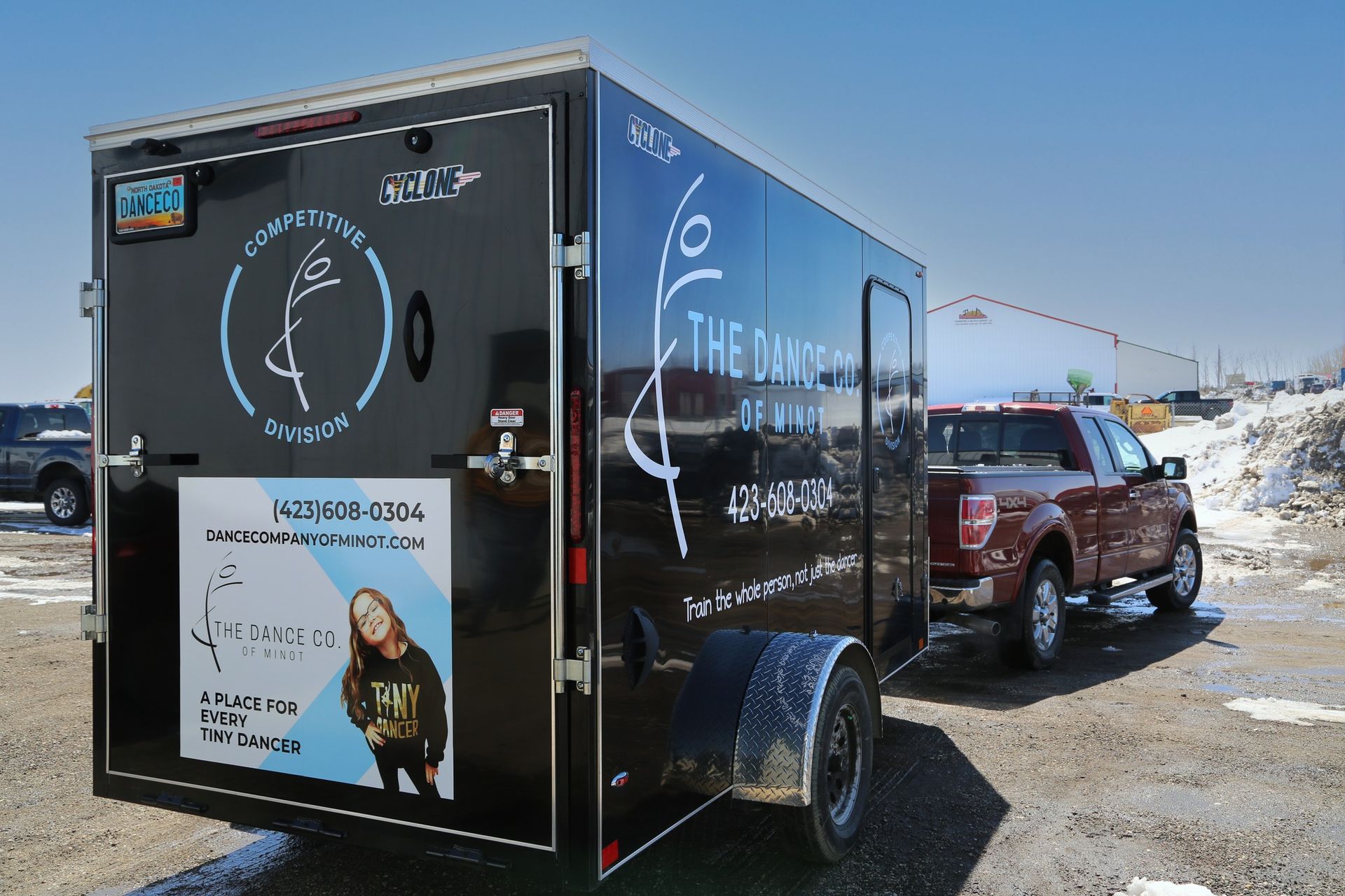 Black trailer with chalk art and a poster parked outdoors on a snowy roadside beside a red pickup truck