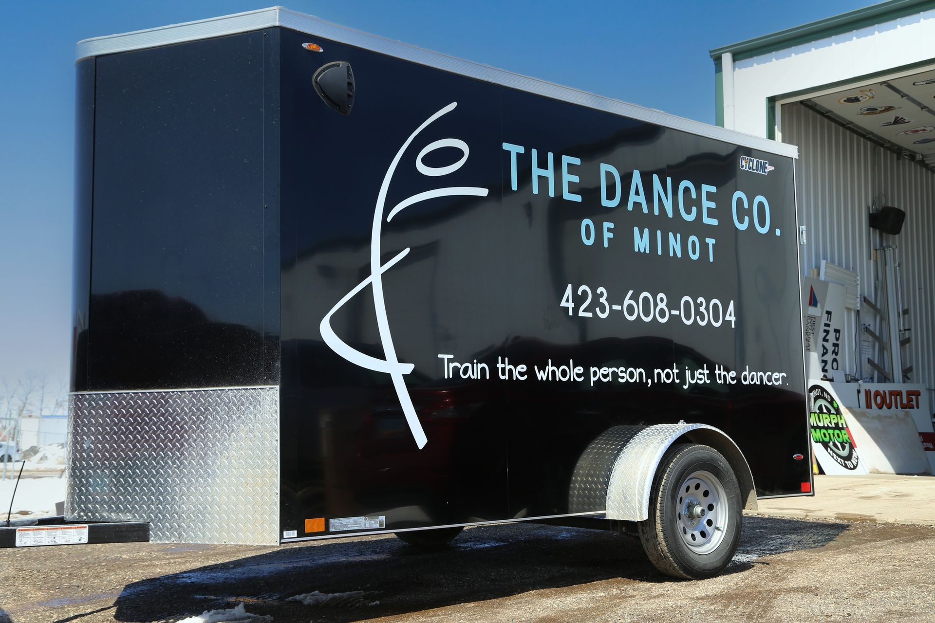 Black trailer for The Dance Co. of Minot with white logo and text, parked outdoors under a blue sky