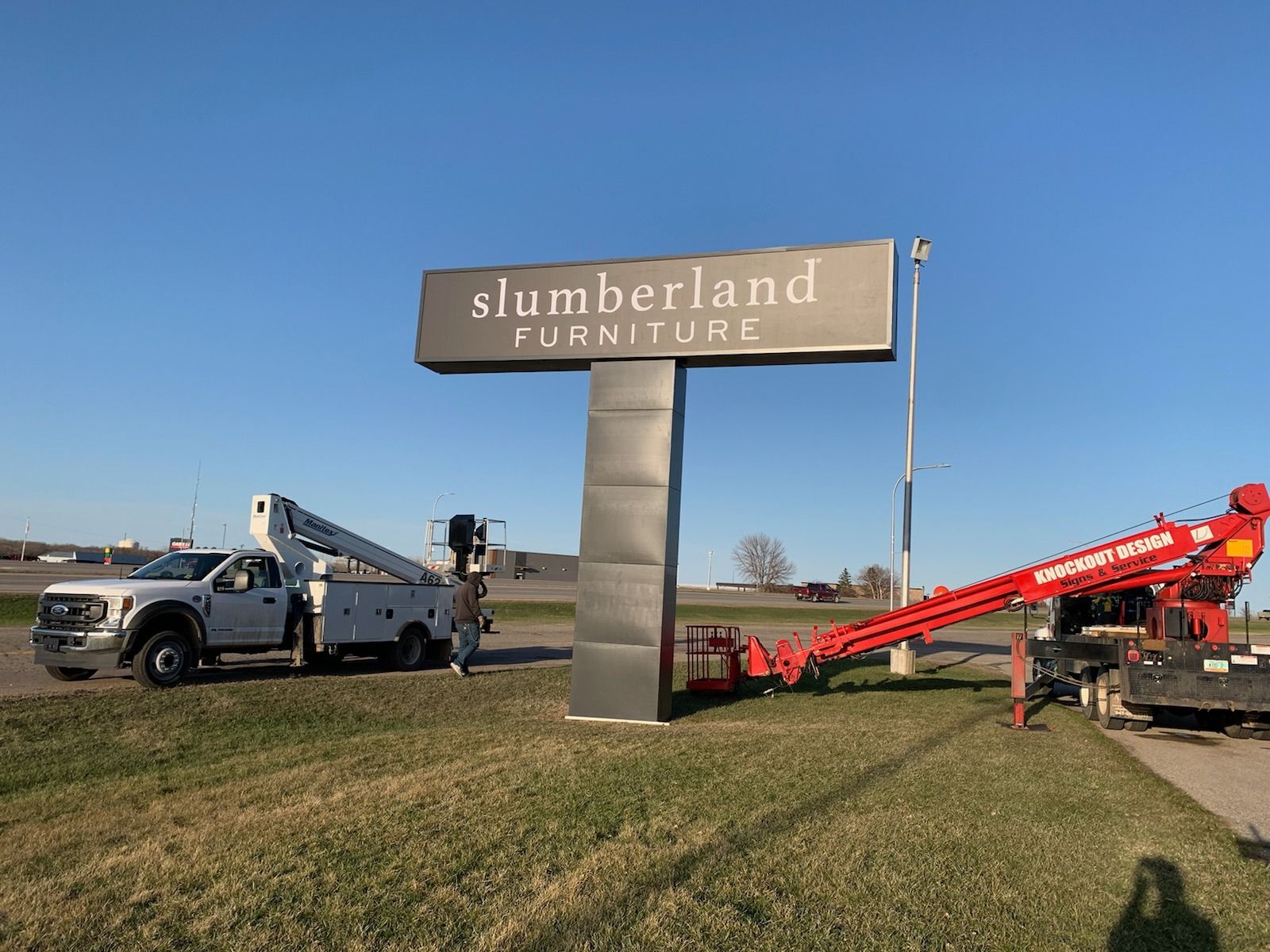 Tow truck beside Slumberland Furniture sign on a grassy roadside under a blue sky