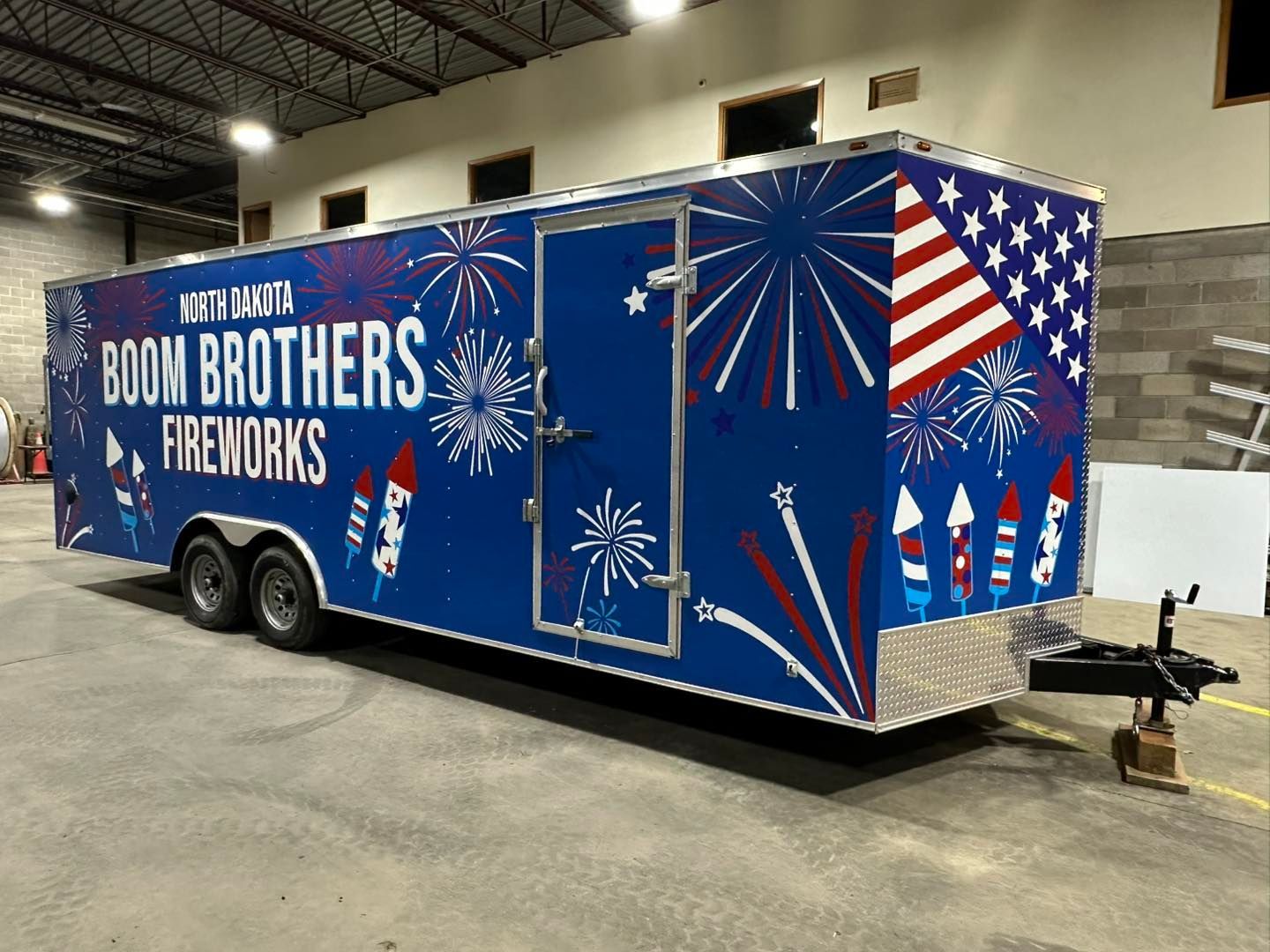 Blue BOOM BROTHERS FIREWORKS trailer with red, white, and blue starburst graphics in a warehouse