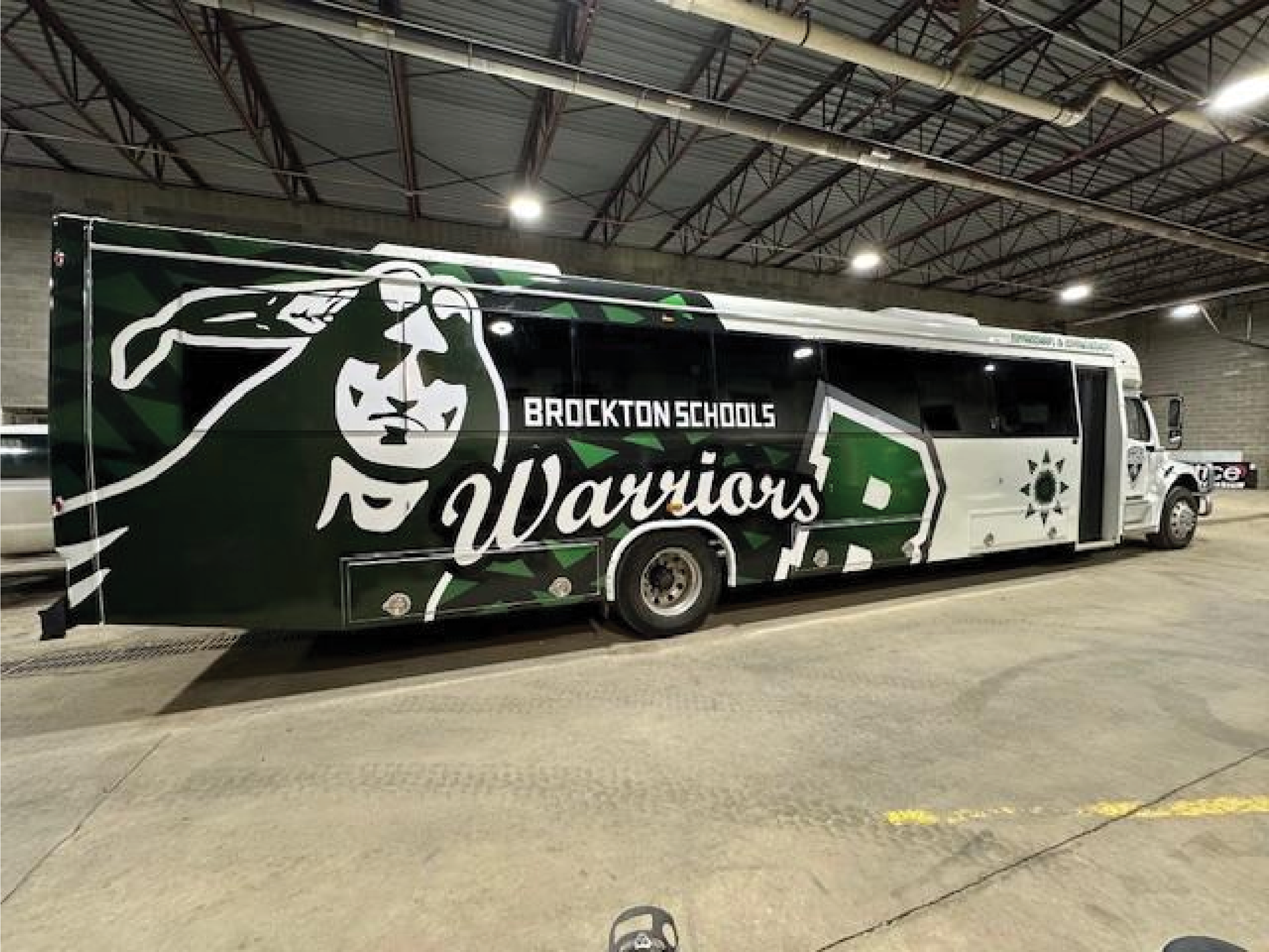 Brockton Schools Warriors team bus parked indoors, black and white with a large warrior graphic.