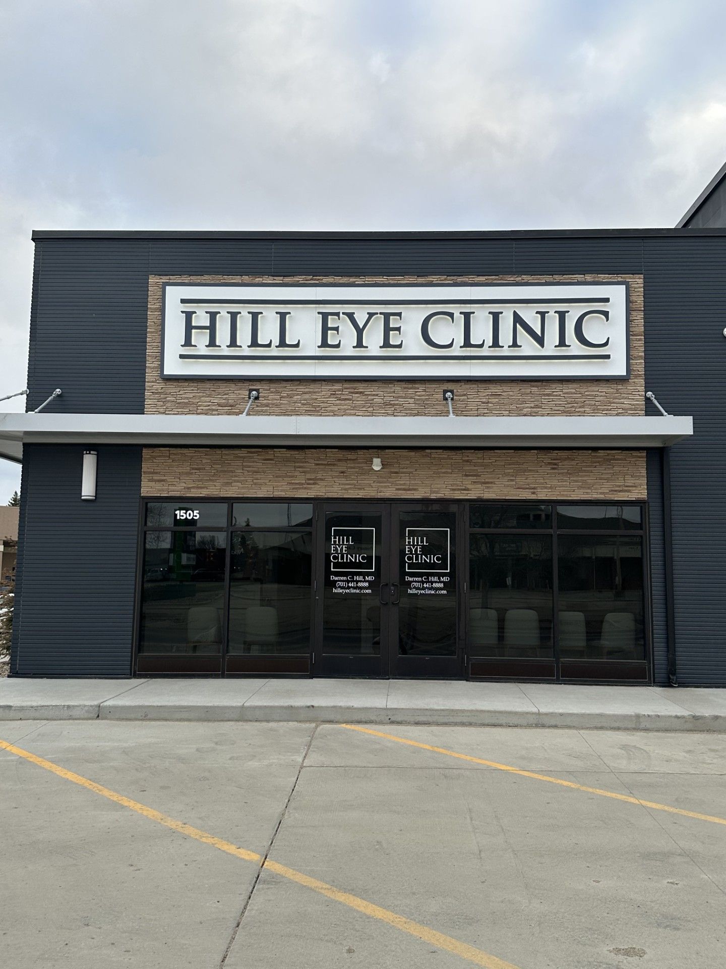 Hill Eye Clinic storefront with dark siding and beige awning under cloudy sky
