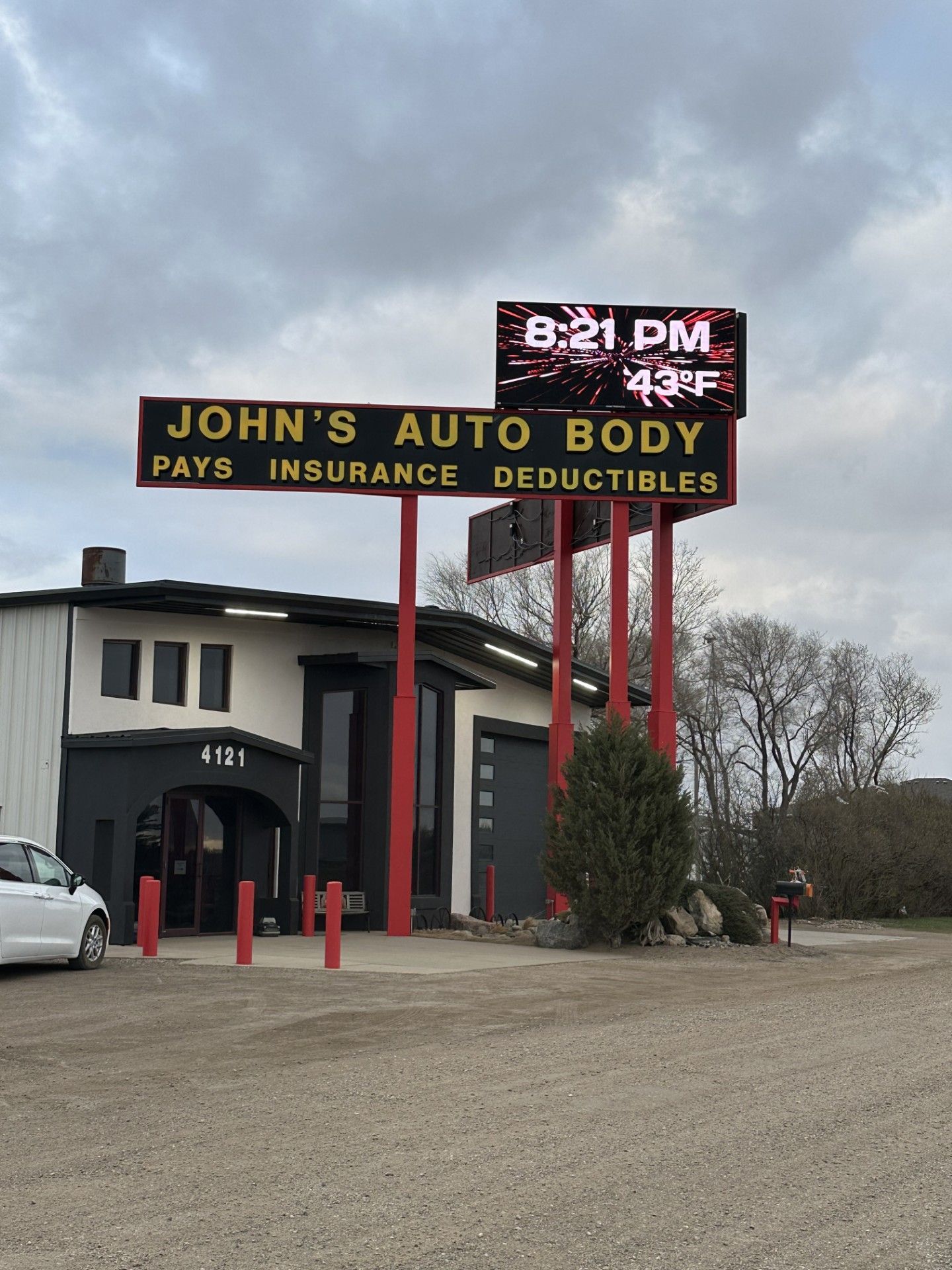 John’s Auto Body sign on red poles beside a dark building on a gravel lot under cloudy skies