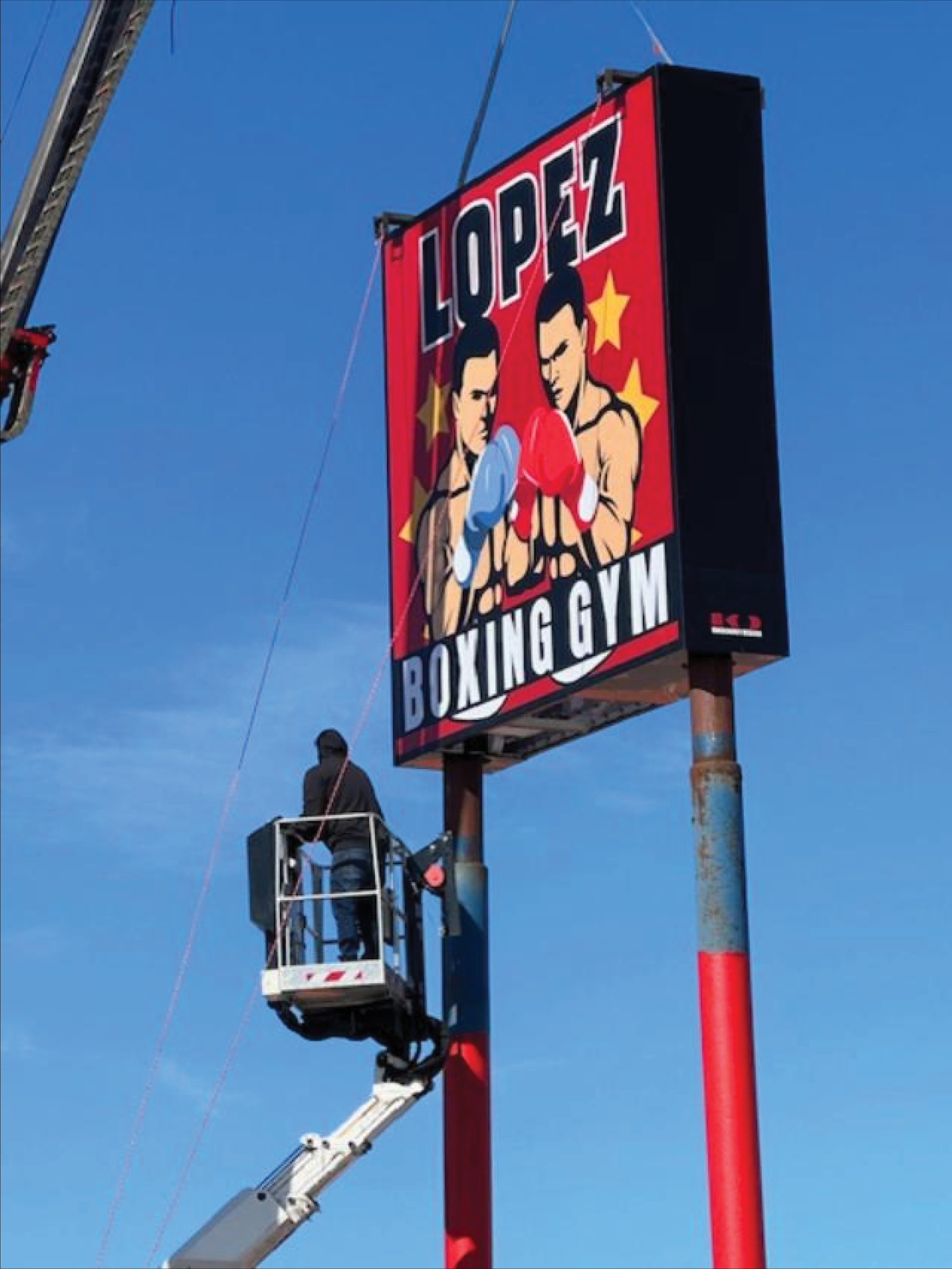 Lopez Boxing Gym sign on a pole against a blue sky, with a worker in a lift below it