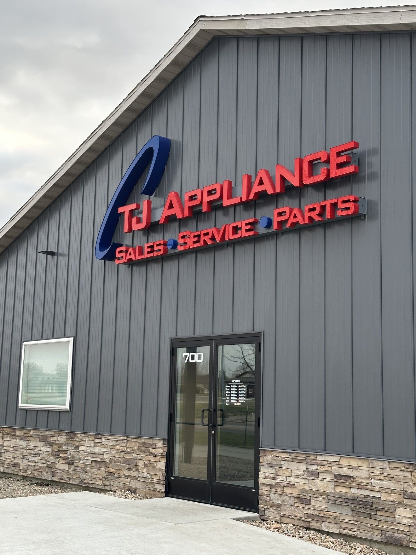 TJ Appliance storefront with gray siding, red sign, and glass entrance under cloudy skies