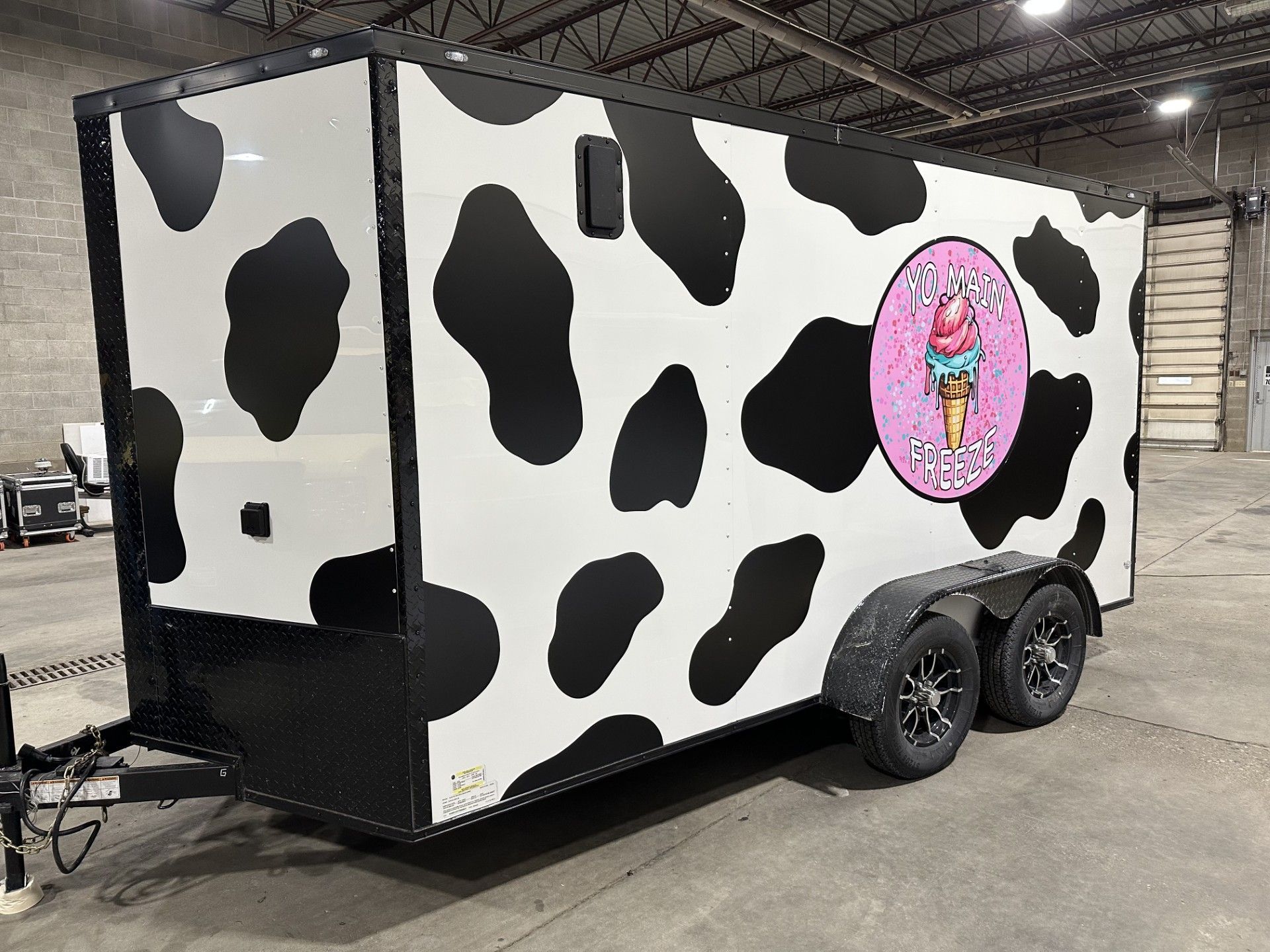 Cow-print trailer with pink logo, parked indoors on a dual-axle chassis